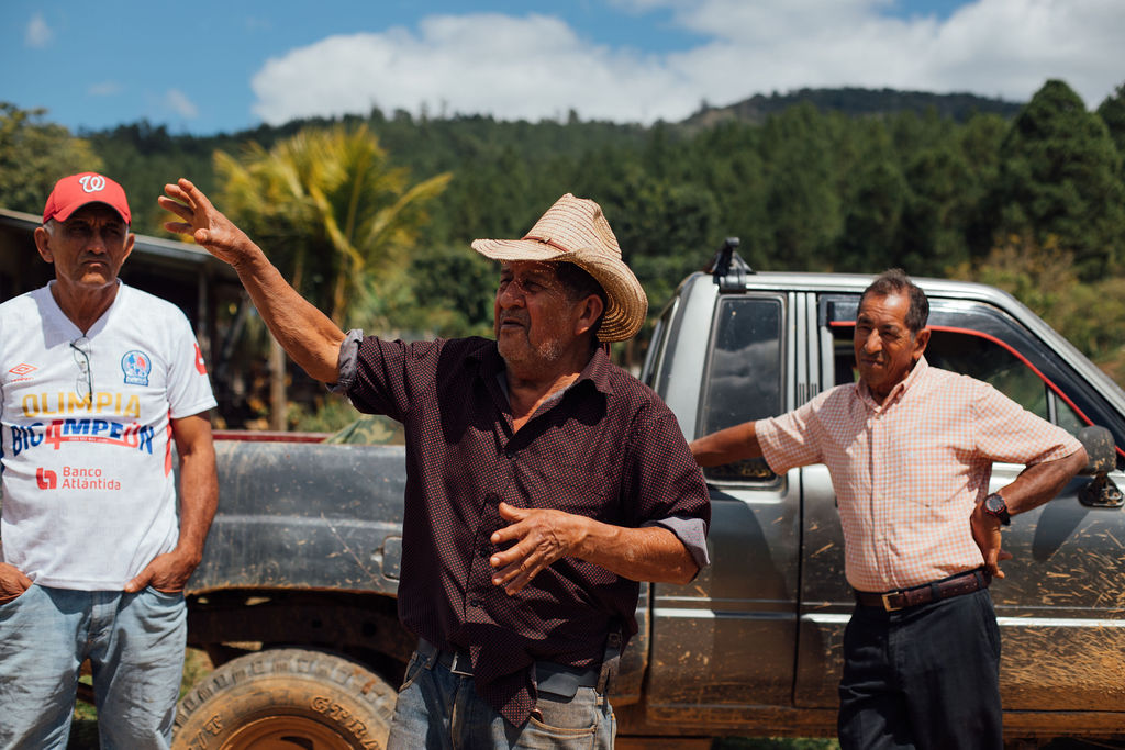 Three men outdoors near a pickup truck, one gesturing with his arm, background of trees and sky