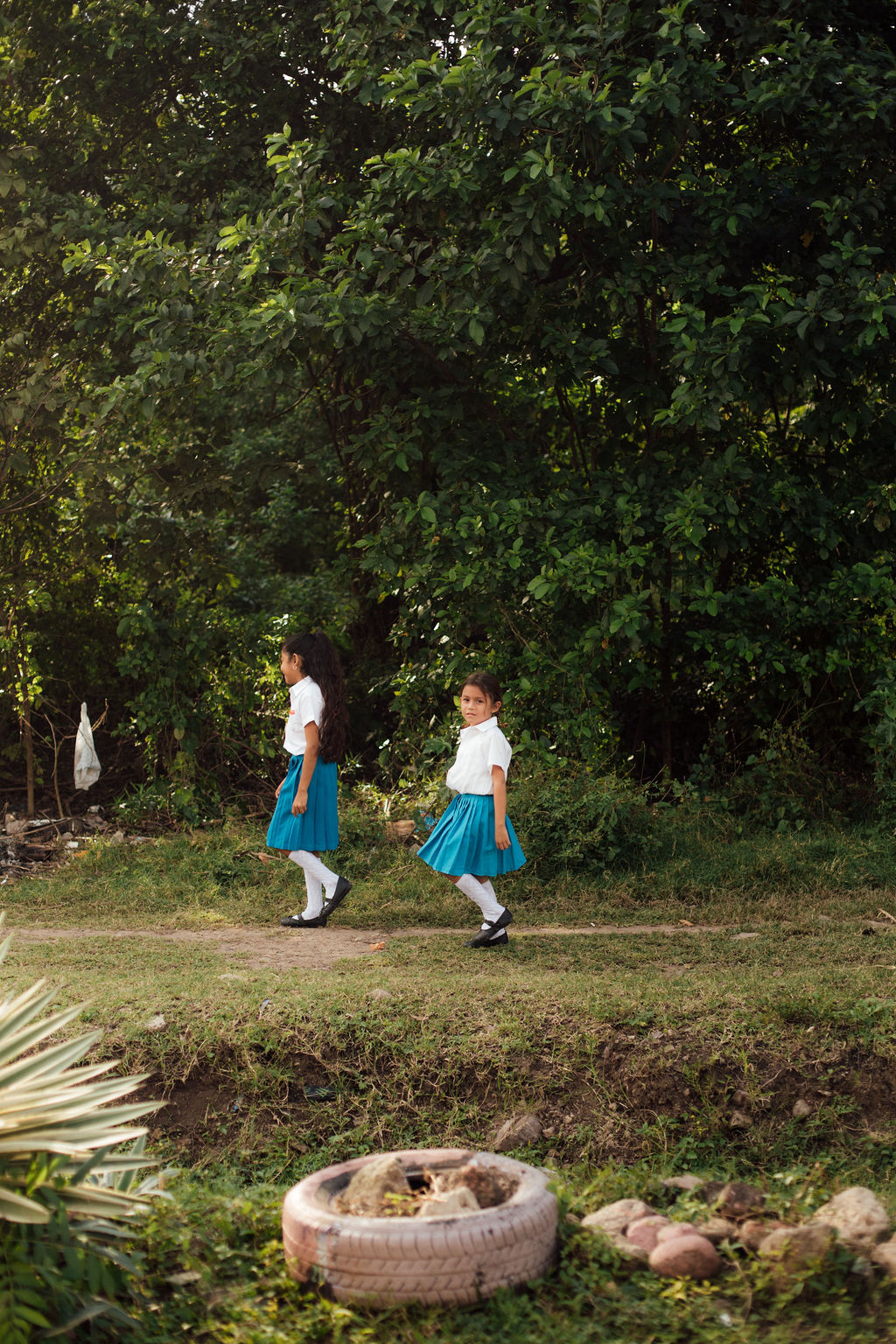 Two children in white shirts and blue skirts walk outdoors near trees, with a grassy area and a tire in the foreground.