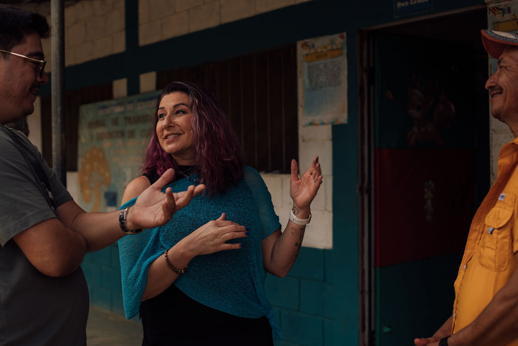 Three people talking and smiling outdoors near a building with posters on the wall.