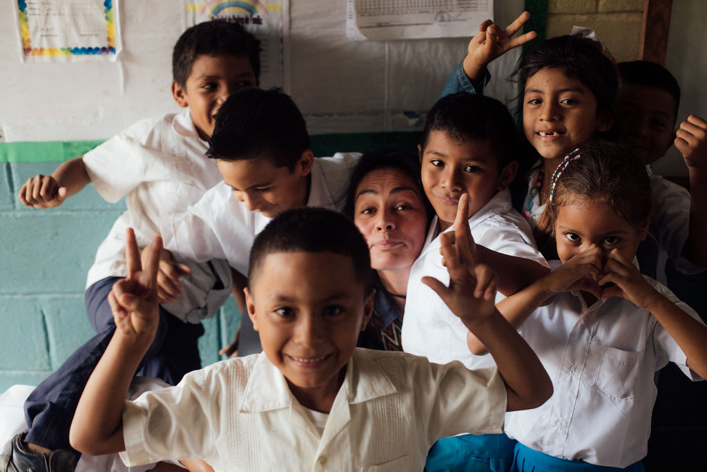 Group of children in white shirts smiling and making gestures in a classroom, some with hands raised or covering faces.