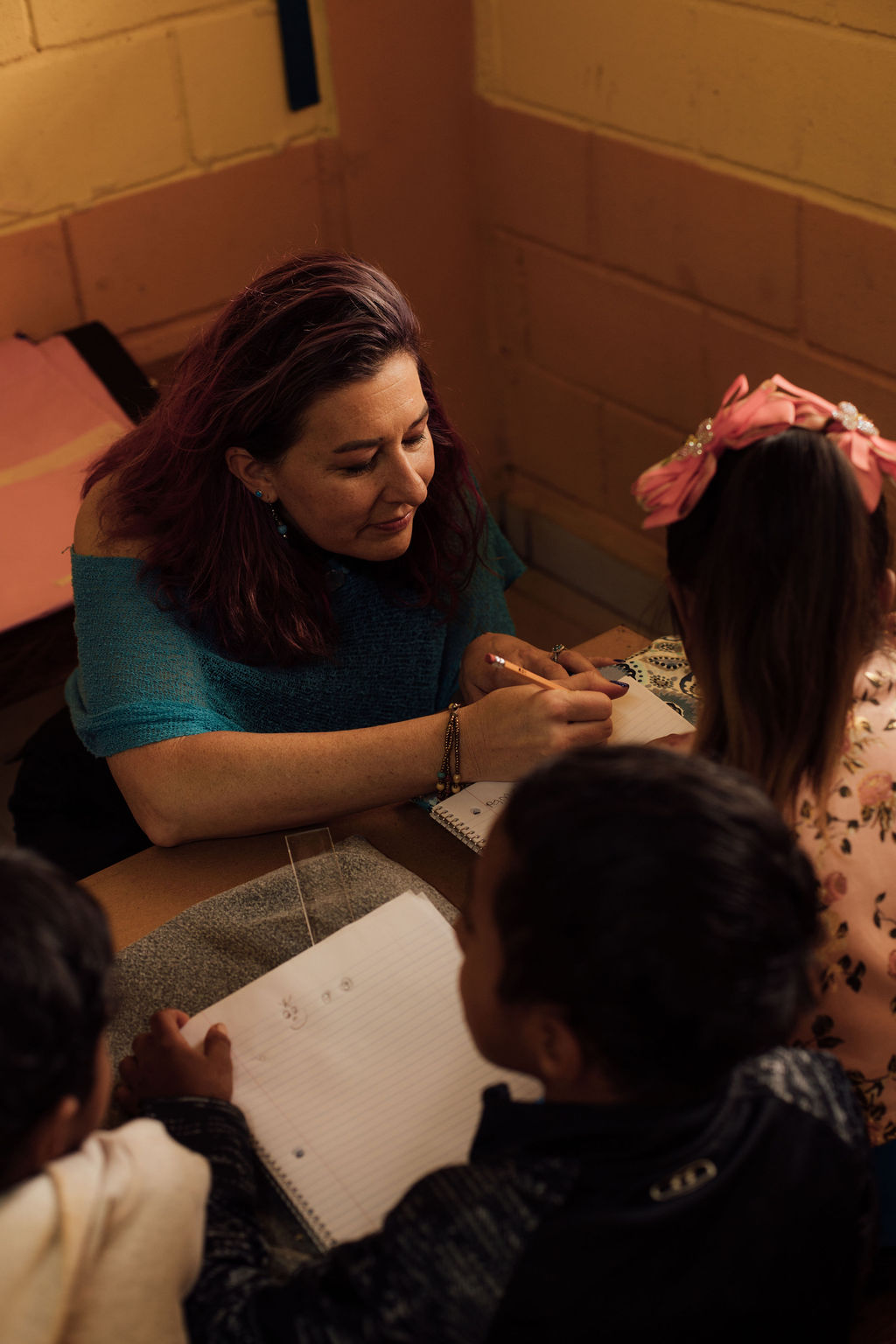 Woman with dark hair writing in a notebook at a table with children, yellow brick wall background.