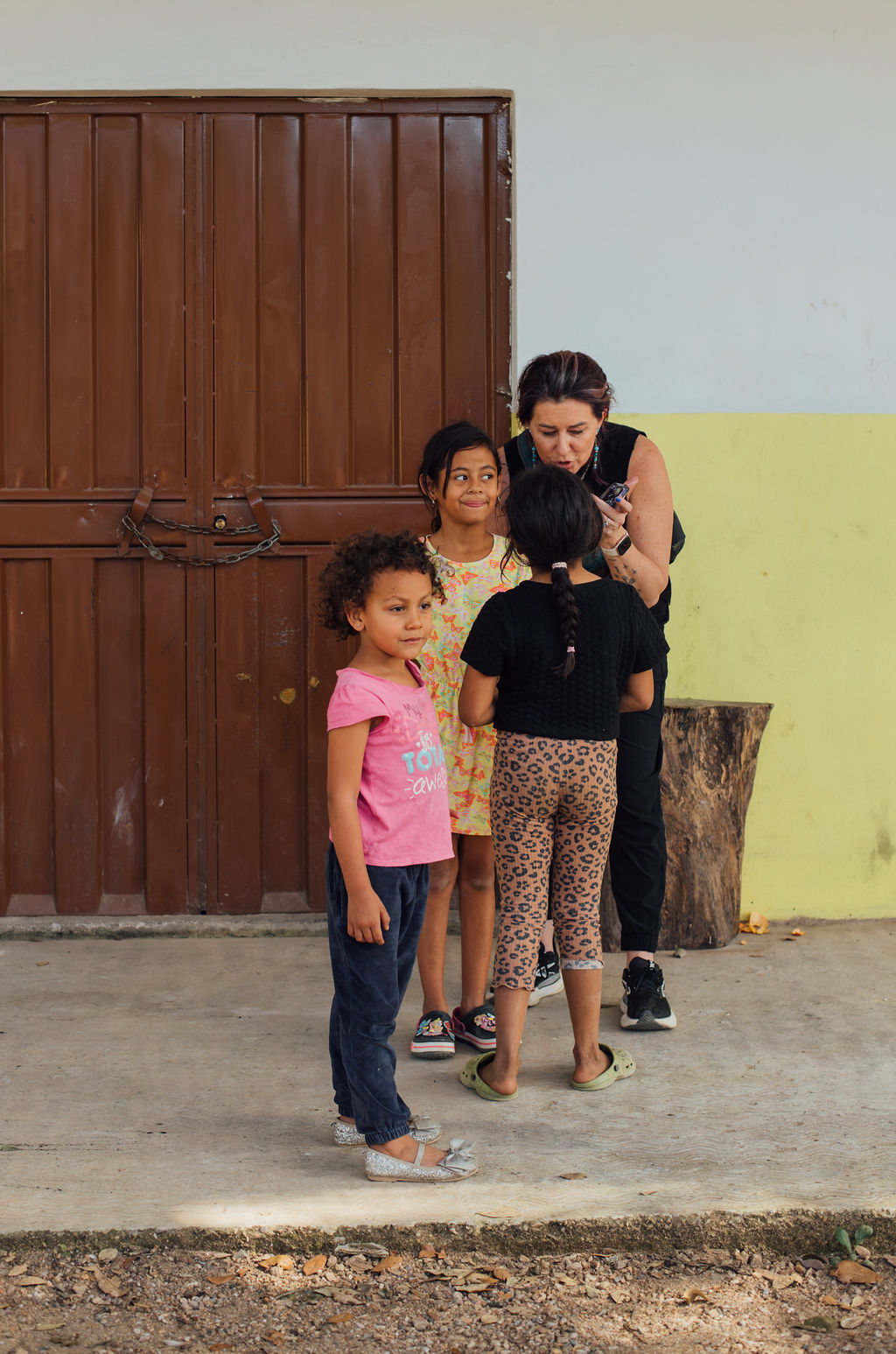 A woman and three children standing outdoors near a wooden door and a yellow wall, engaged in conversation.