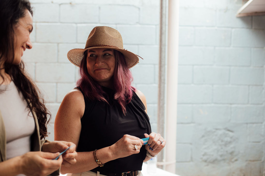 Two women sitting indoors, one wearing a hat and black top, the other with long dark hair, smiling and holding objects.
