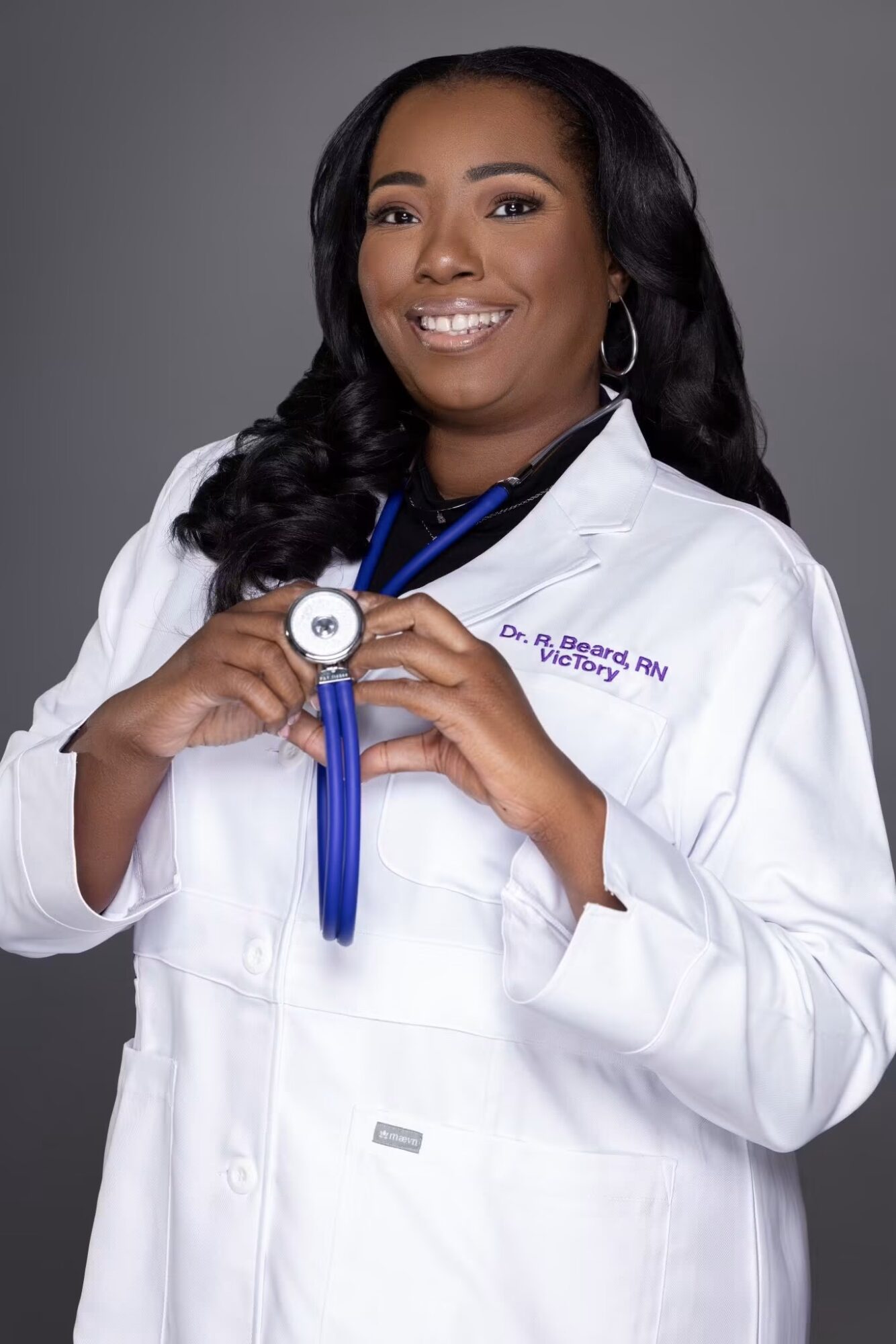 Female doctor in white coat holding a stethoscope, smiling, against gray background.