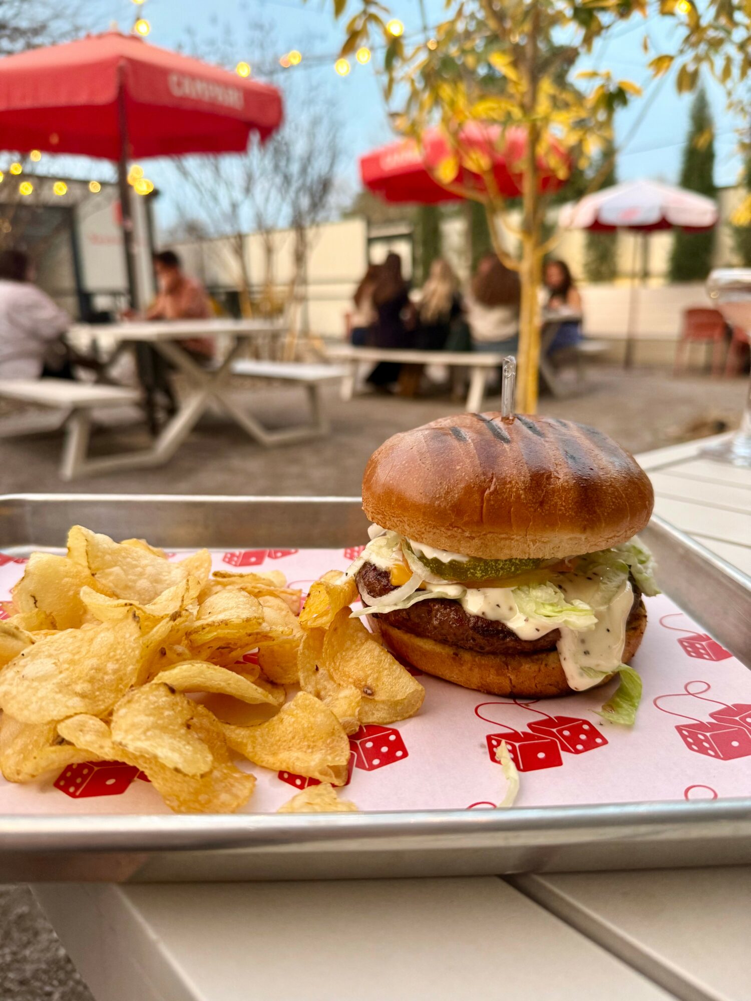 Burger with lettuce and sauce, side of potato chips on tray, outdoor patio with tables and umbrellas, people sitting in background.