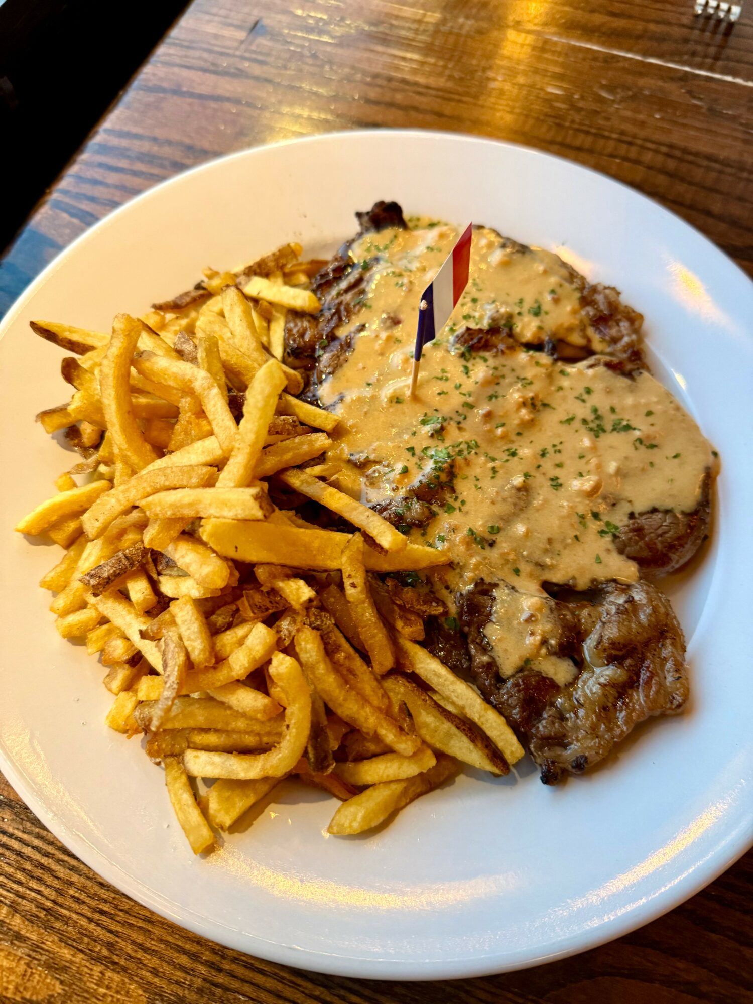 Plate with French fries and a meat dish topped with gravy, garnished with herbs, on a wooden table.