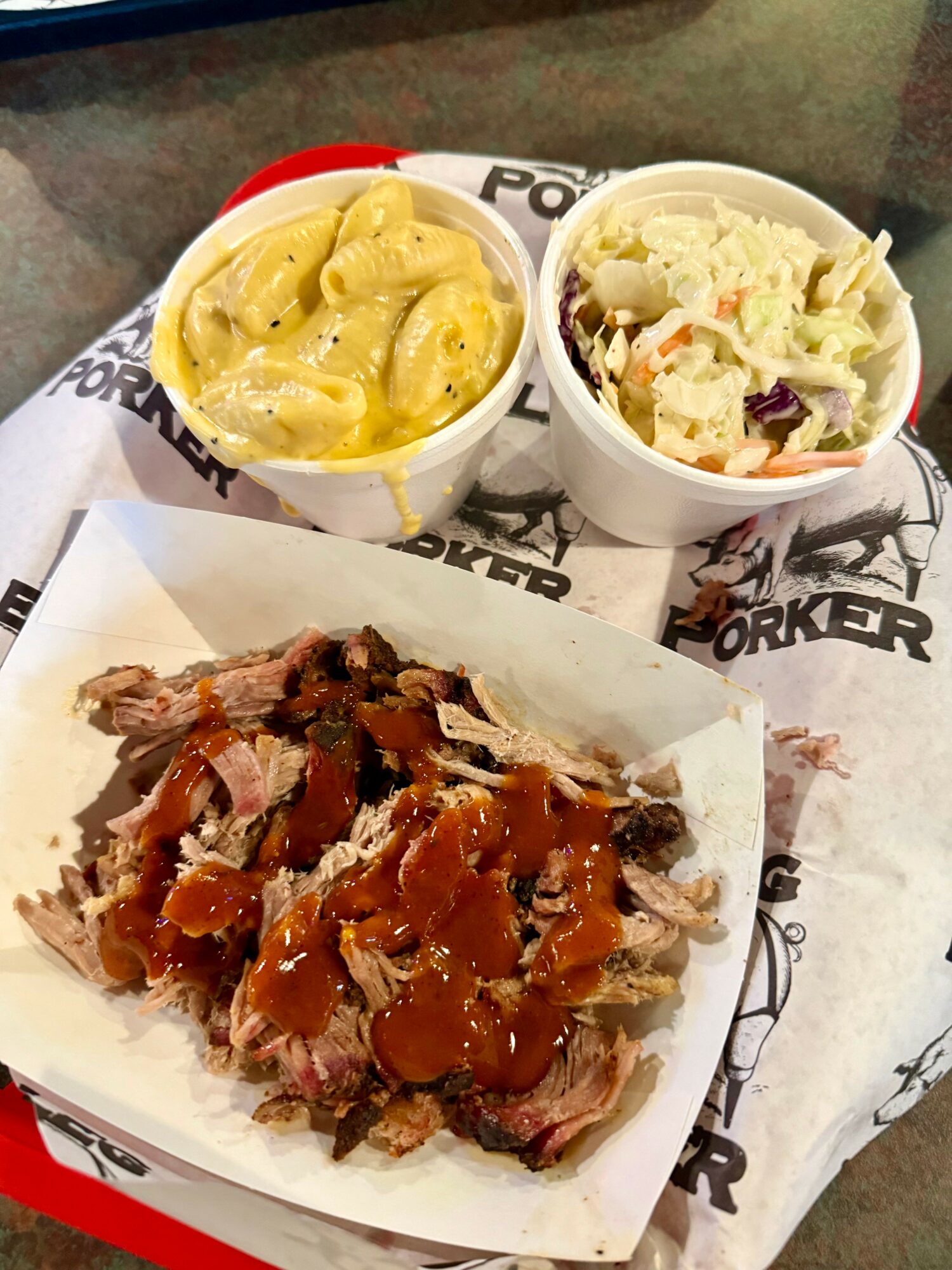 Two bowls of side dishes and a plate of pulled pork with barbecue sauce on paper, on a tray.