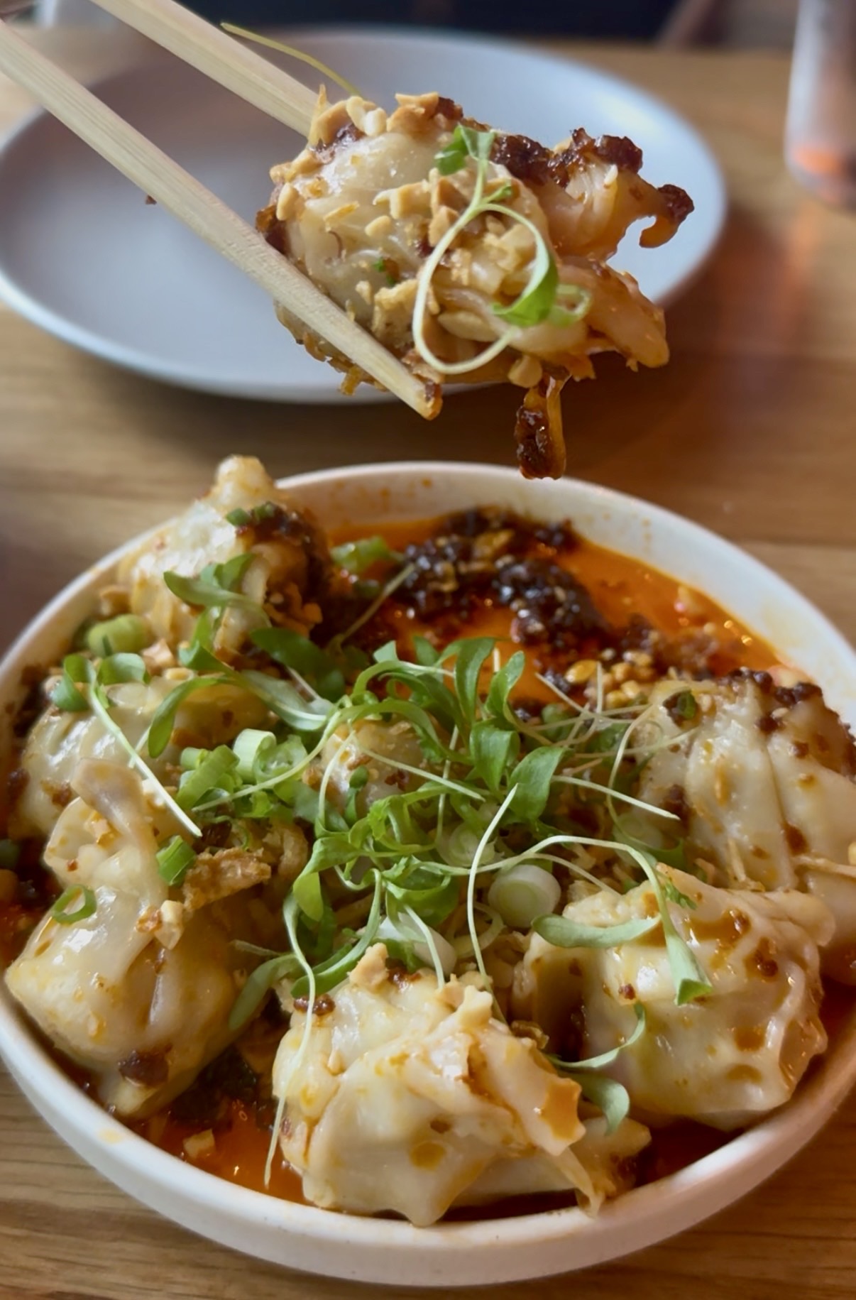Dumplings in a spicy red sauce topped with green sprouts, with a piece held by chopsticks above a white bowl.