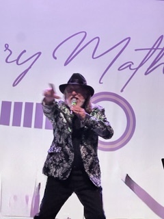 Person wearing a hat and patterned jacket pointing forward on stage with a backdrop.