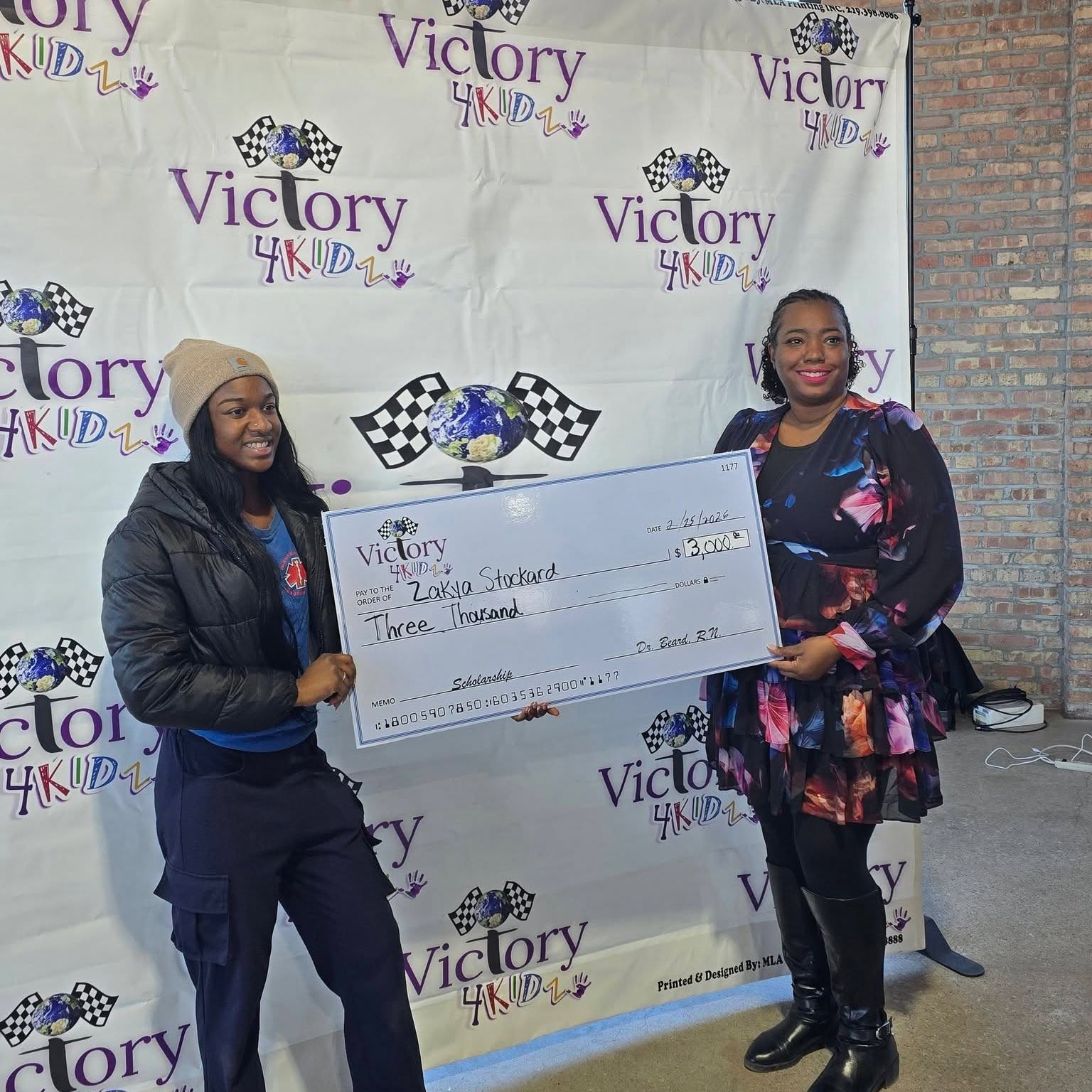Two women holding a large check in front of a Victory 4KIDz backdrop, smiling. One wears a beanie and jacket, the other a colorful dress.