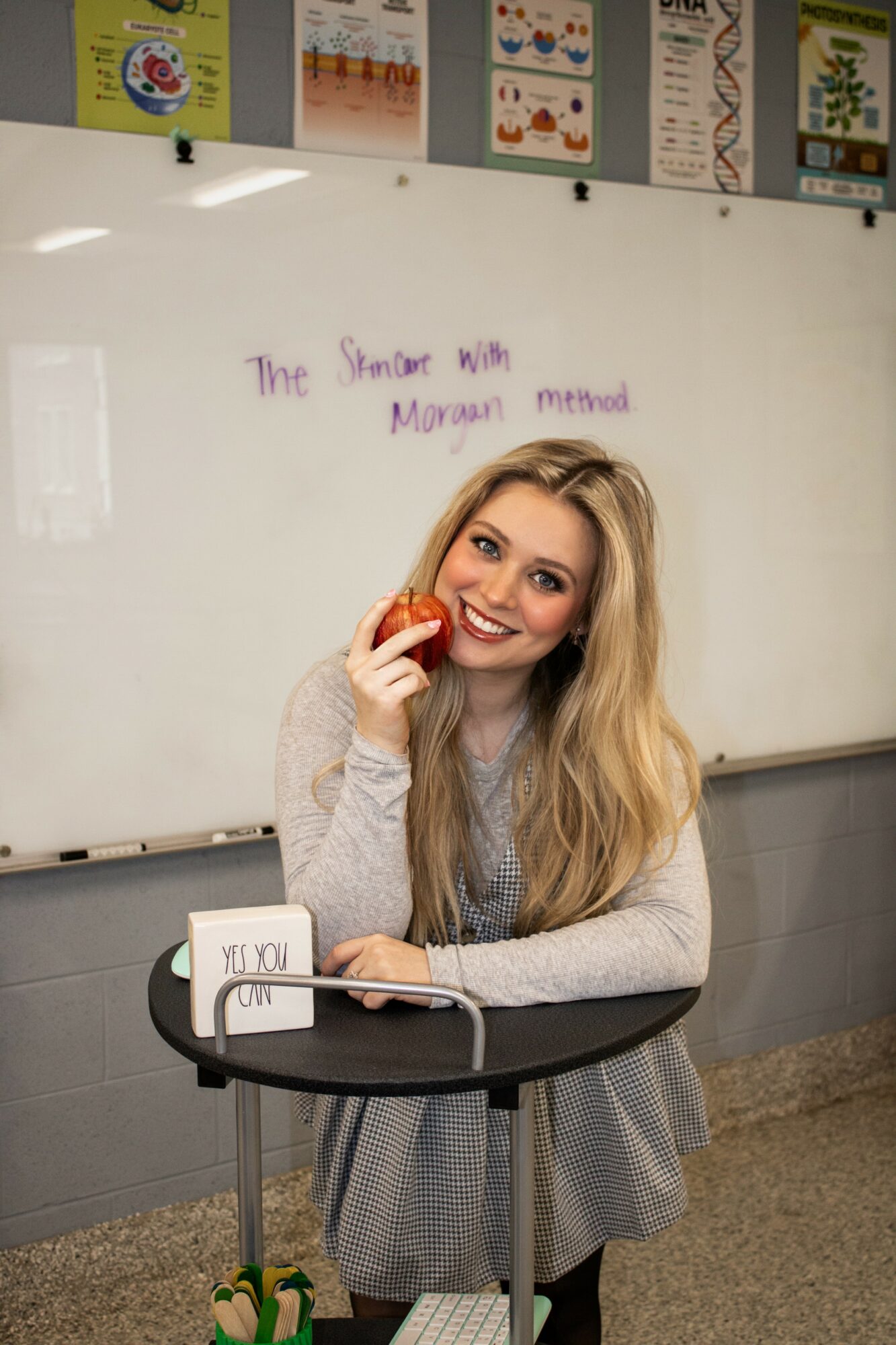 Smiling woman with long blonde hair holding an apple in a classroom, whiteboard with writing behind her.