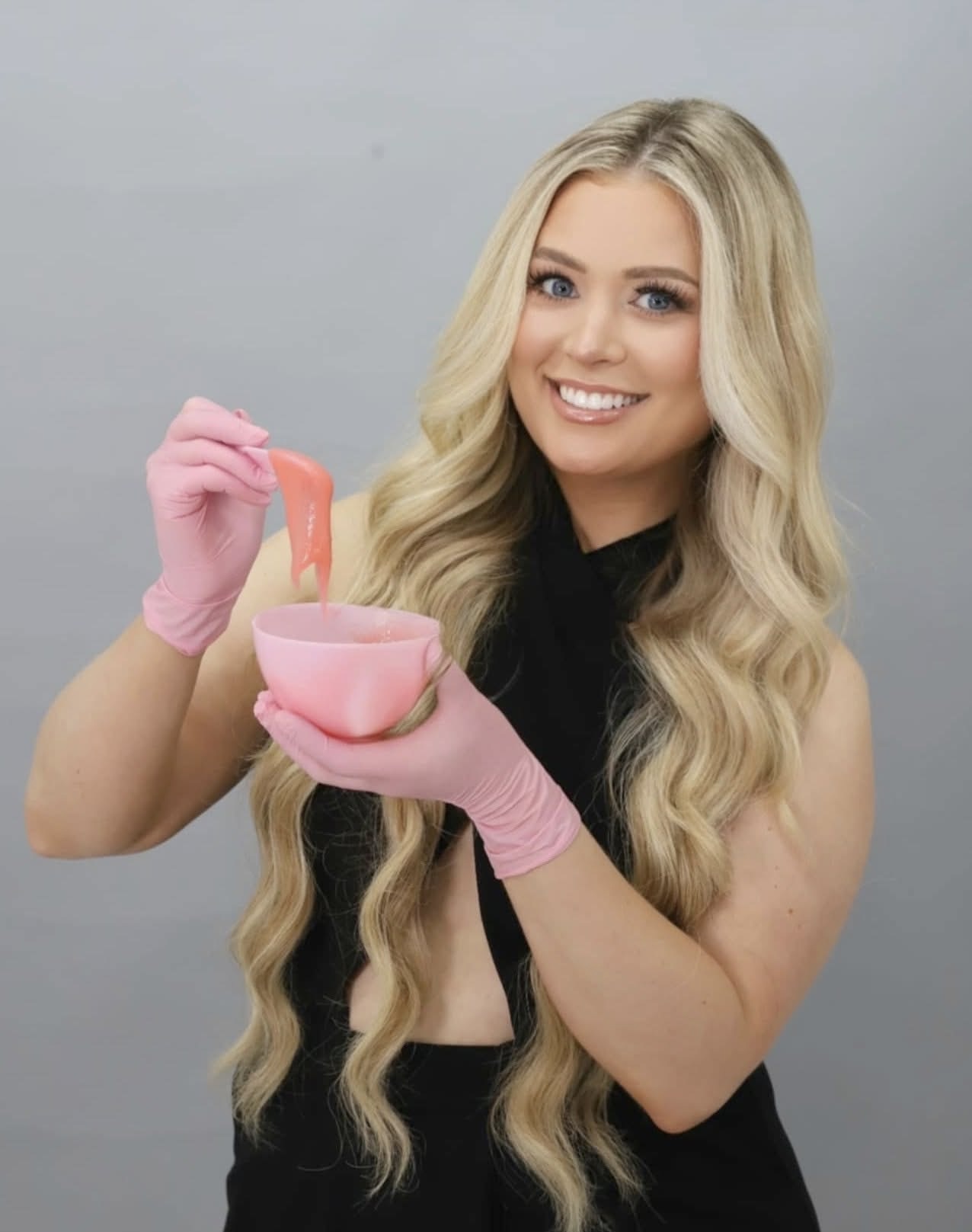 Young woman with long blonde hair smiling, wearing pink gloves, holding a pink bowl and a spatula, against a plain background.