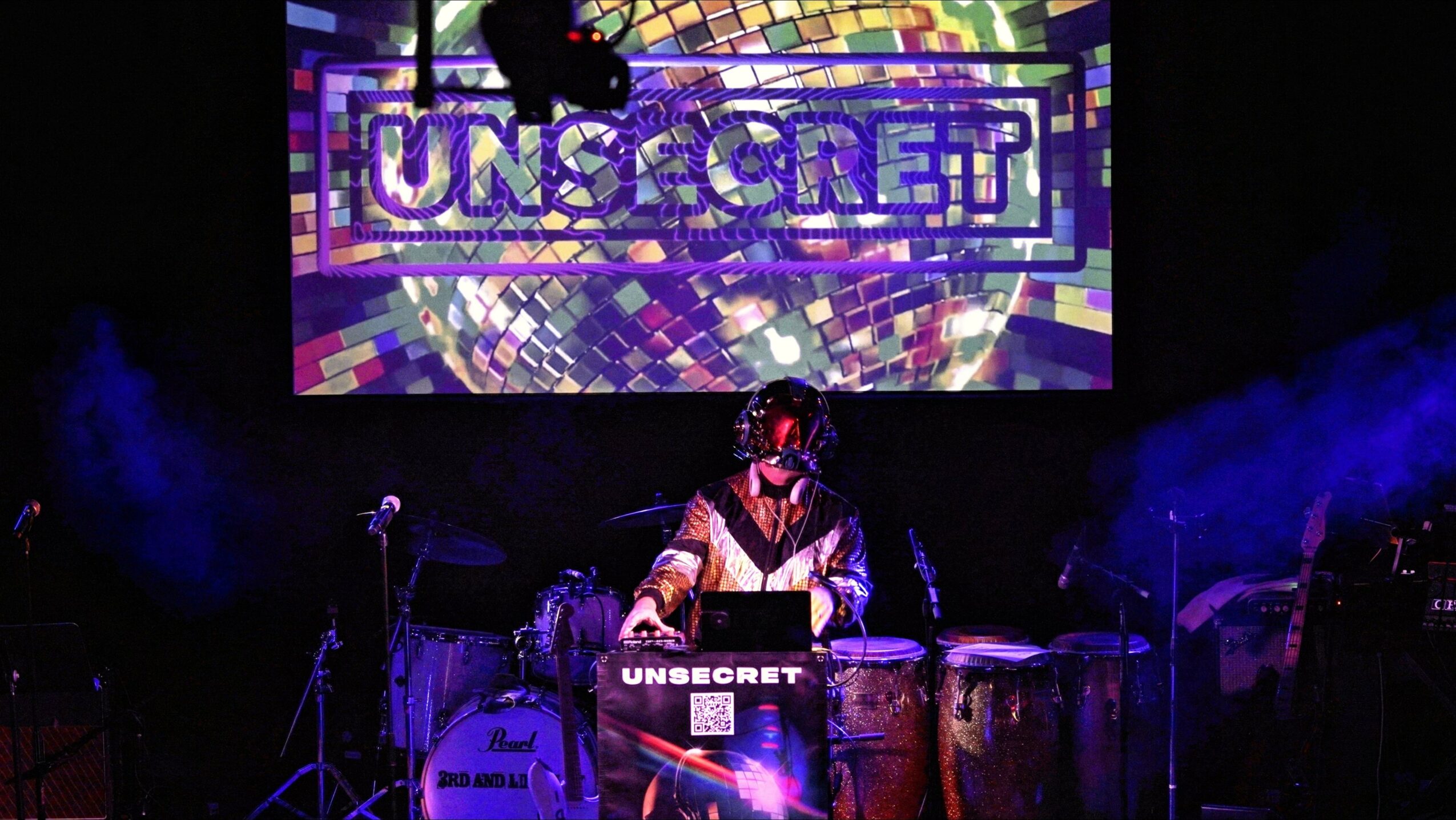 Performer playing drums on stage with a large screen displaying 'UNSECRET' behind them.