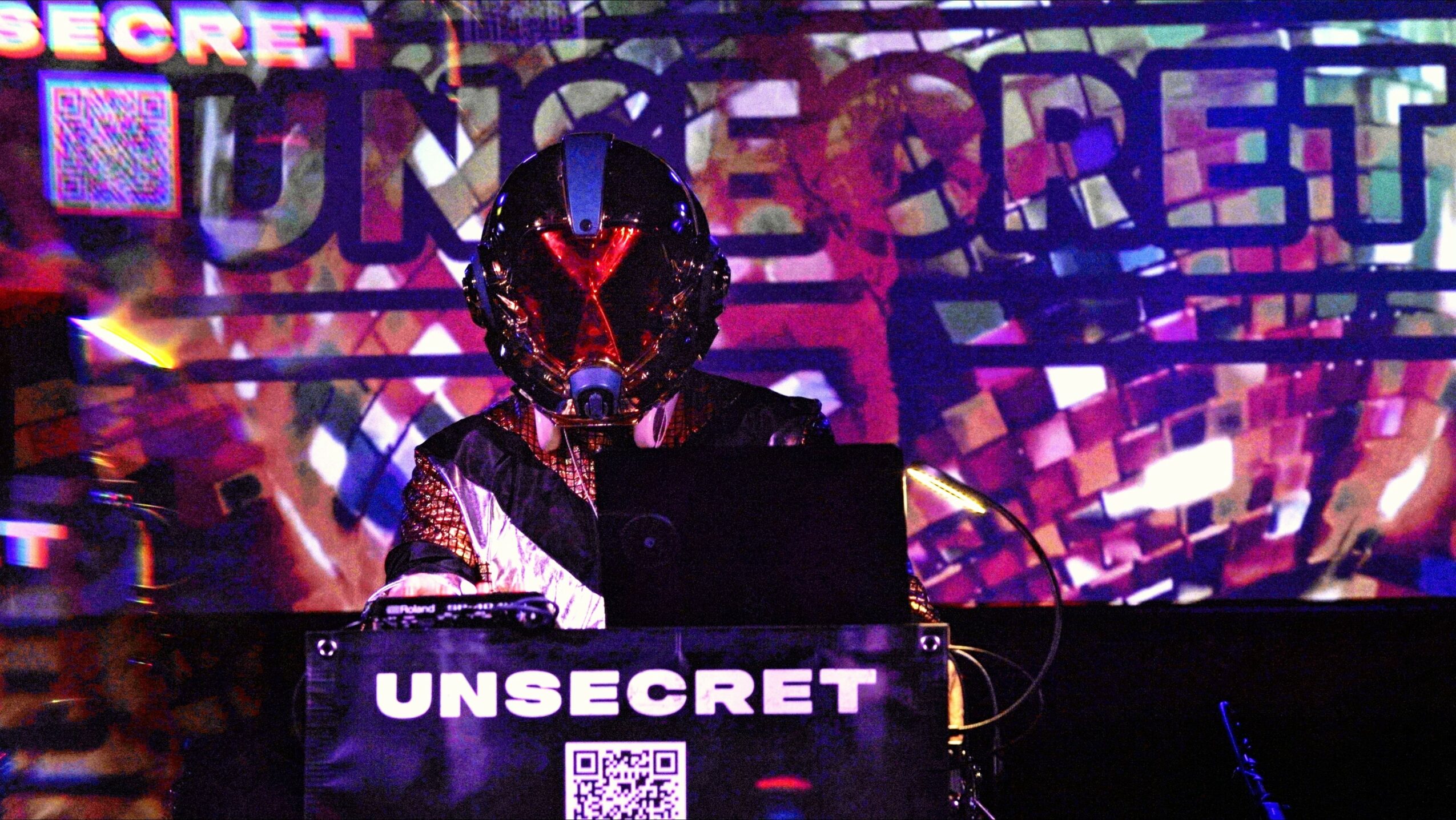 Person wearing a mask with a dark background and colorful lights, standing behind a sign that says 'UNSECRET'.