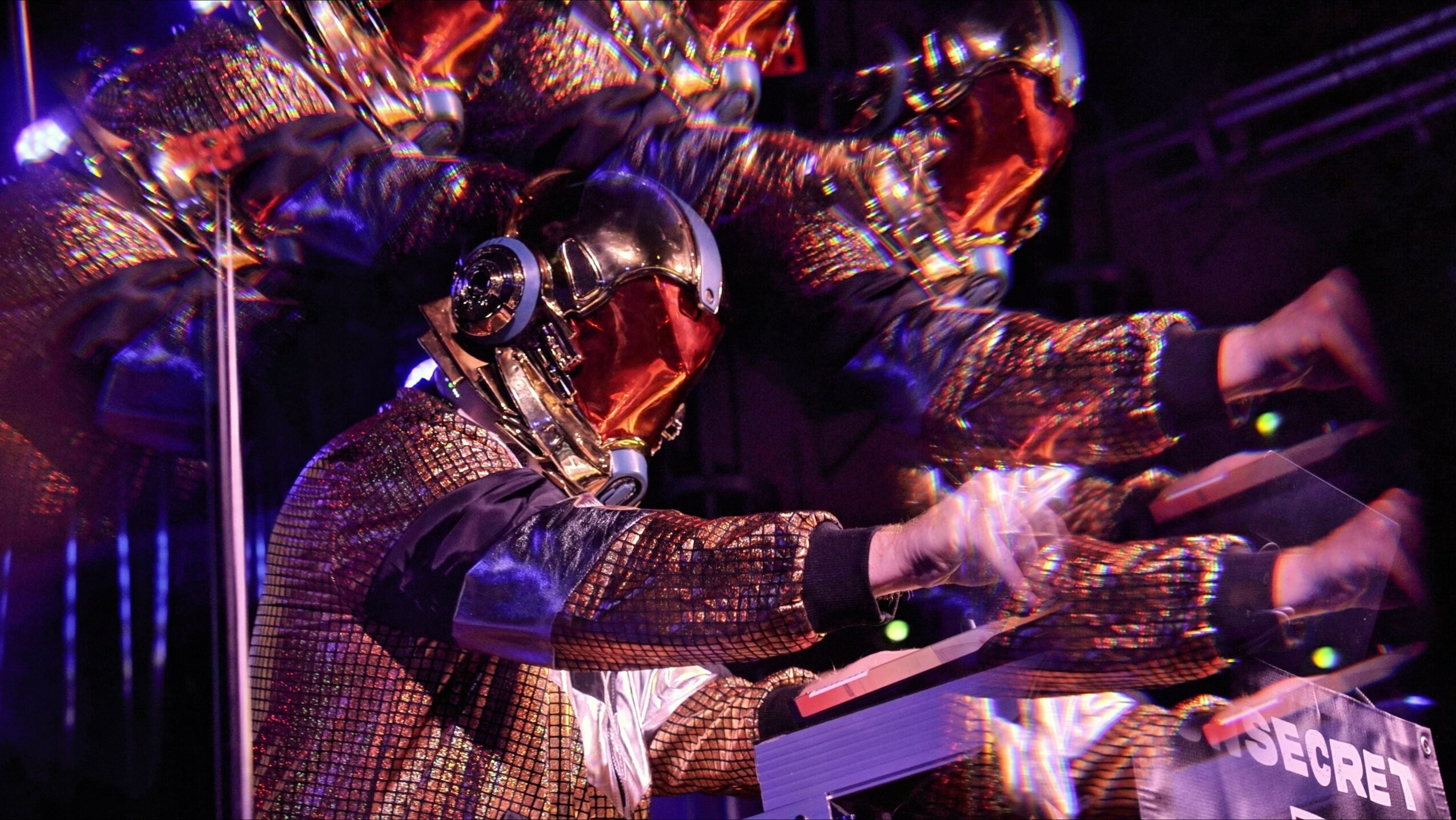 Group of people wearing reflective helmets and patterned jackets, reaching towards electronic equipment in a dark setting.
