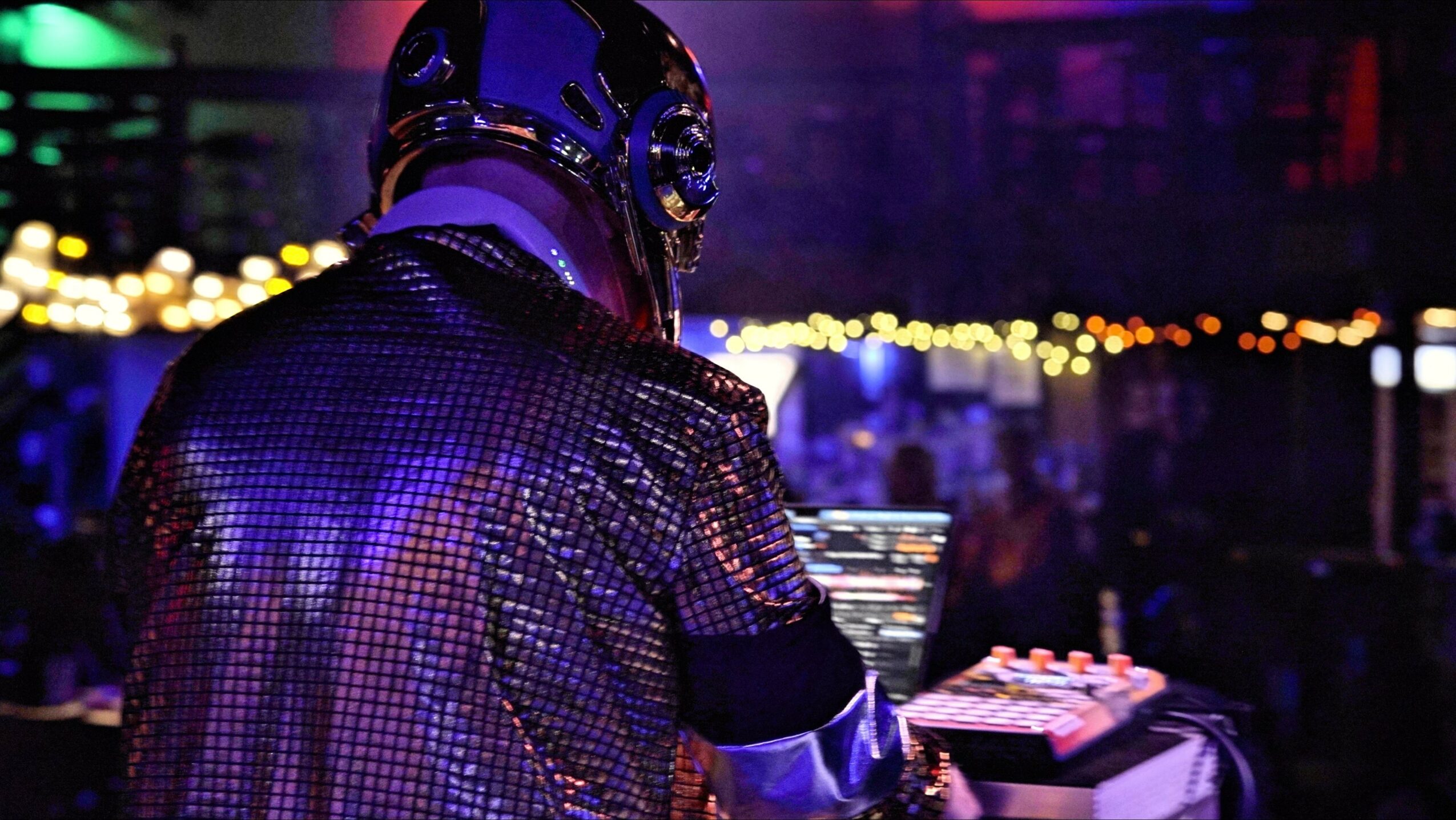Person wearing a helmet and shiny jacket DJing at a nightclub with colorful lights in the background.