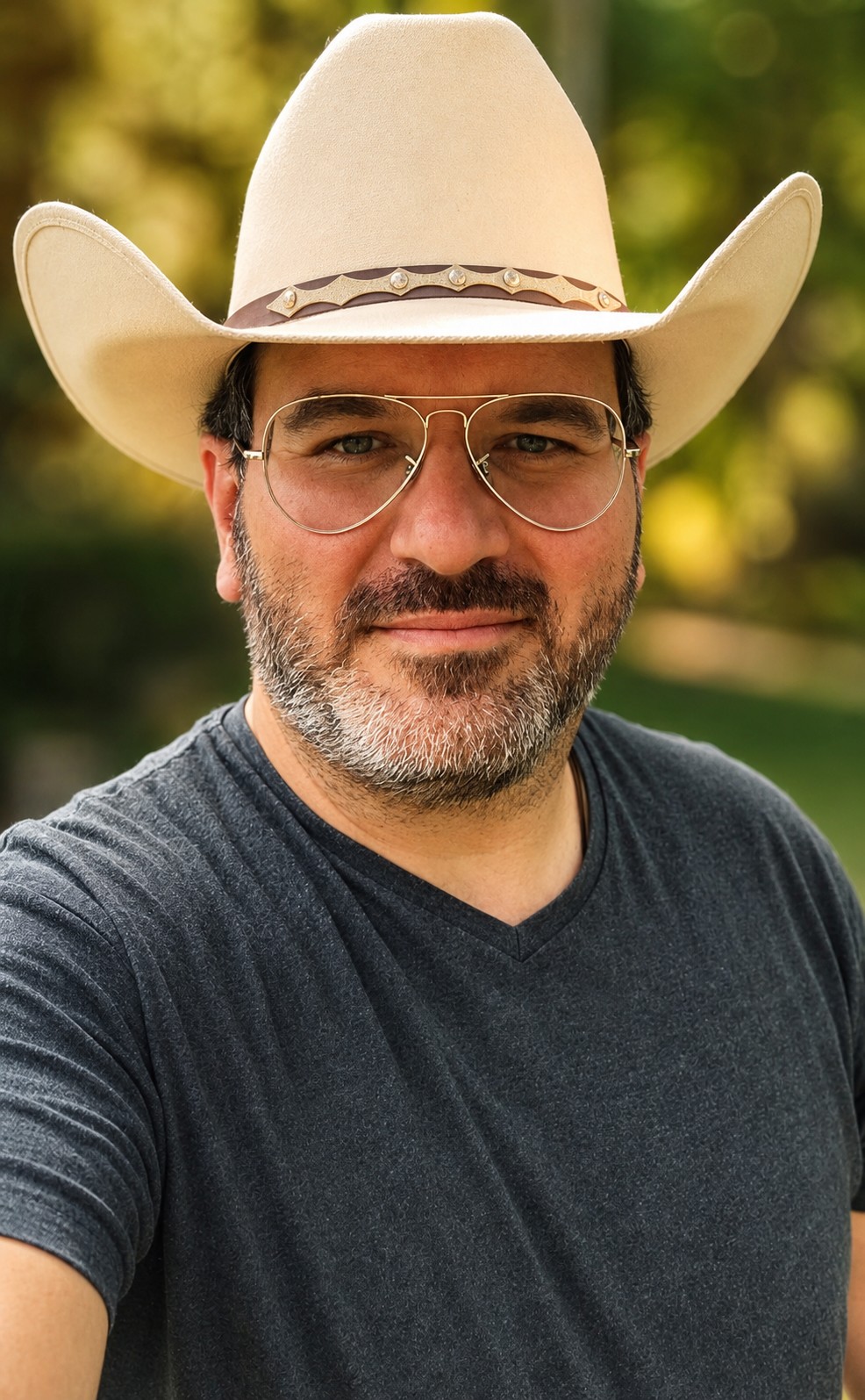 Man wearing a cowboy hat and glasses, outdoors with blurred green background.