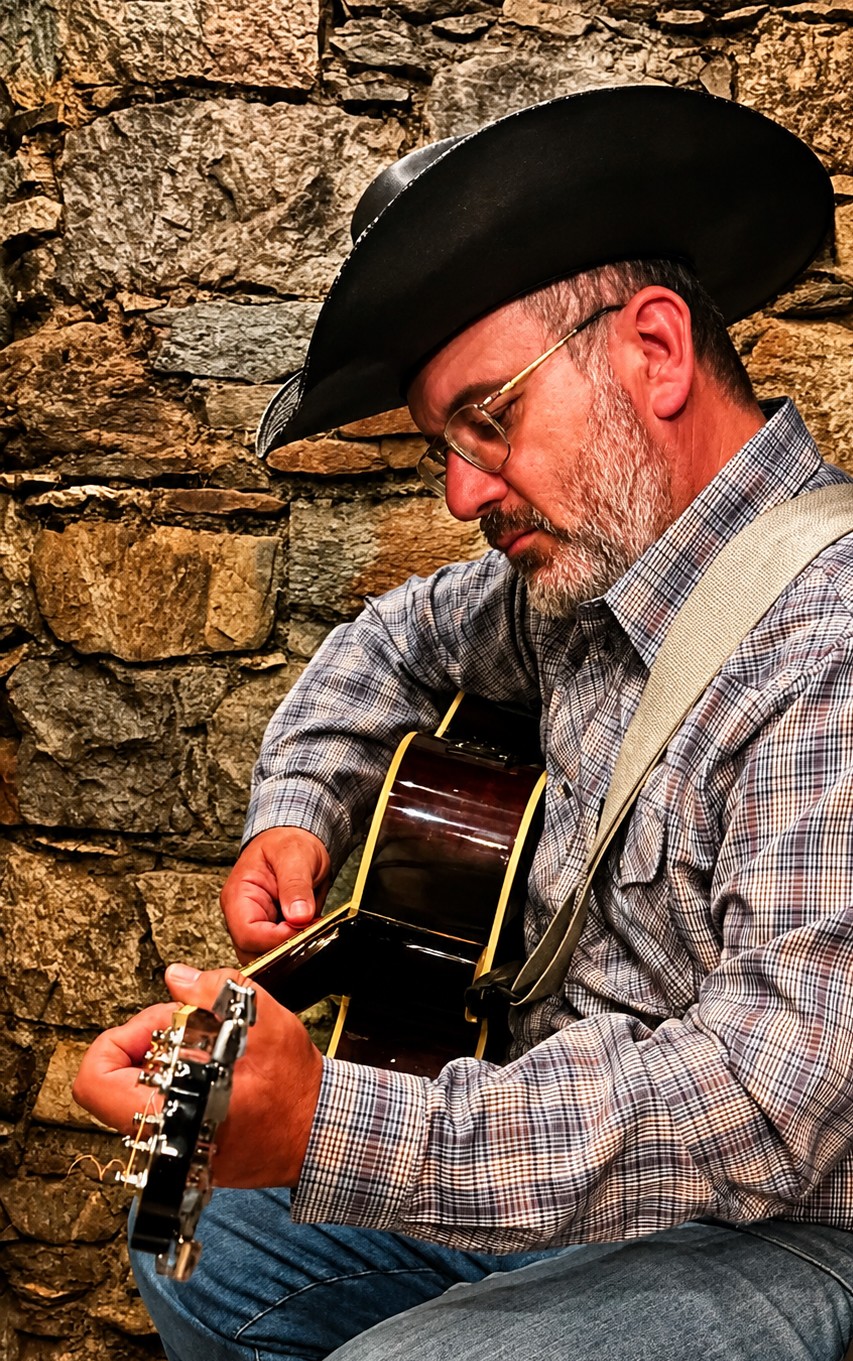 Man wearing a cowboy hat and glasses playing an acoustic guitar against a stone wall background.