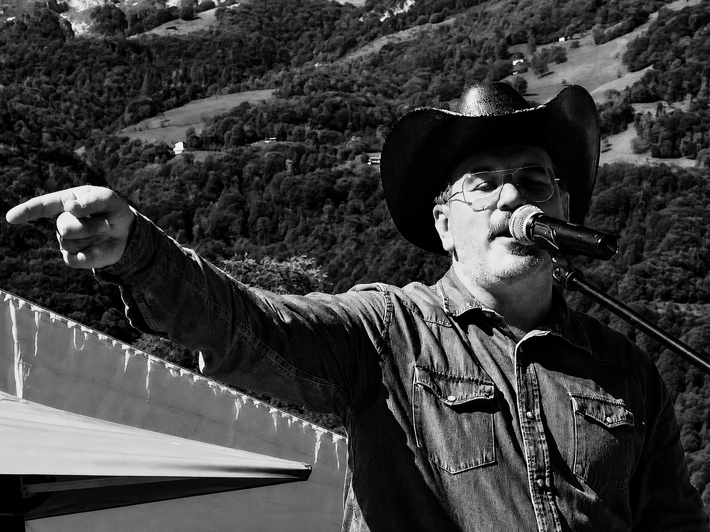 Man wearing a cowboy hat and glasses speaking into a microphone, pointing to the left, outdoors with hills in background.