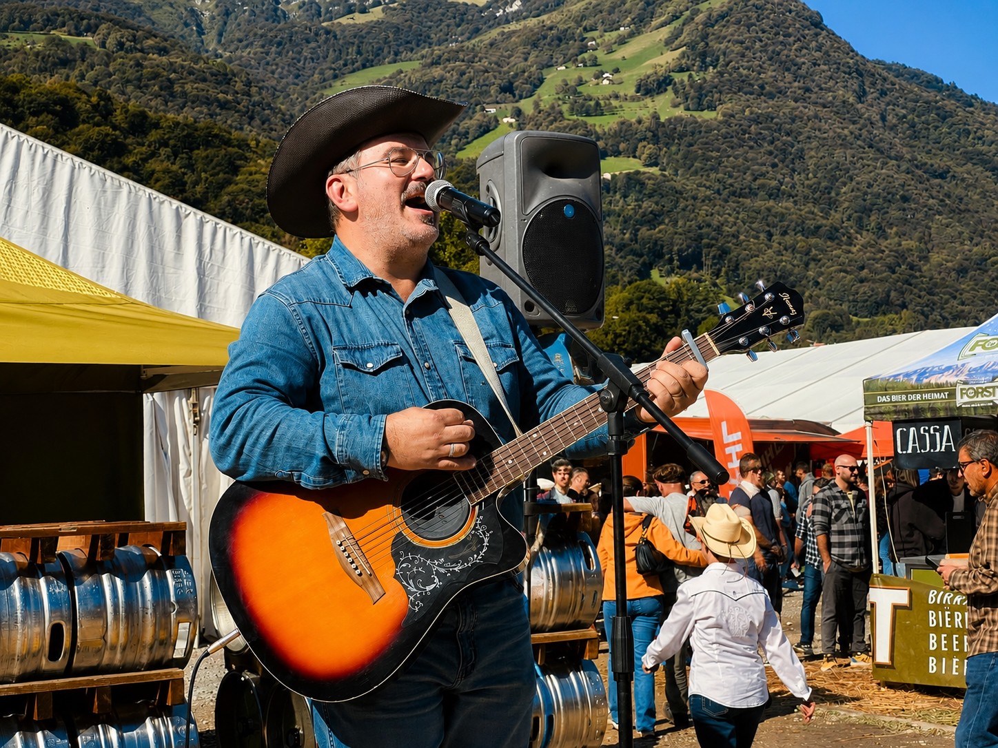 Man wearing a cowboy hat playing guitar and singing outdoors with tents and people in the background.