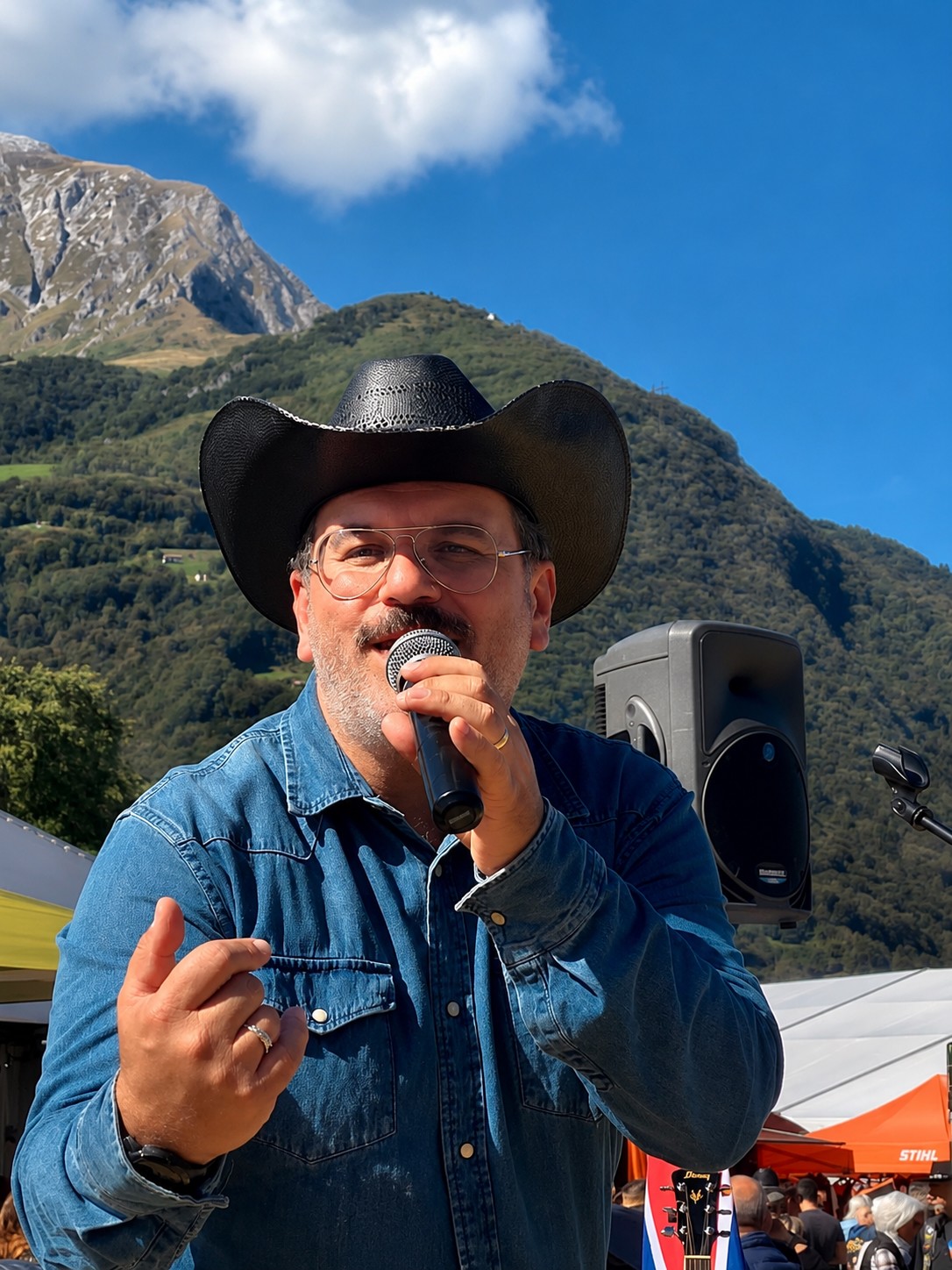 Man wearing a cowboy hat and glasses speaking into a microphone outdoors with mountains and blue sky in background.