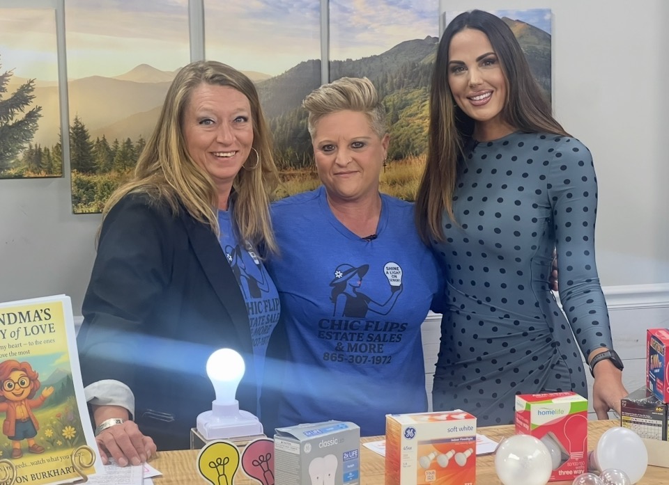 Three women standing behind a table with colorful boxes and a lit candle, smiling indoors with a scenic mountain view in the background.