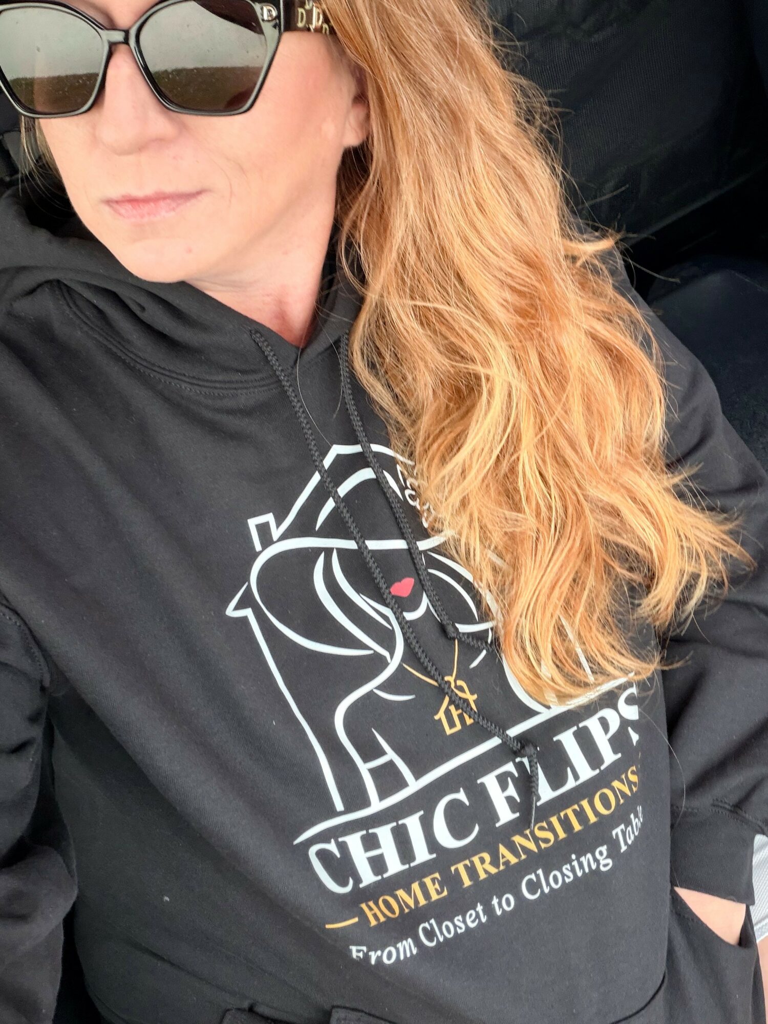 Woman with long wavy hair wearing sunglasses and a black hoodie with a lion logo, sitting in a car.