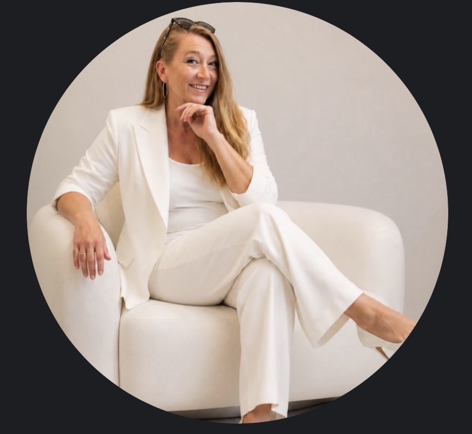 Woman in white suit sitting on a white sofa, smiling and resting her chin on her hand.