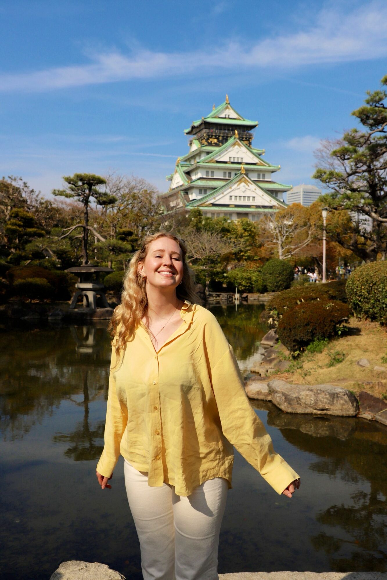 Woman with blonde hair in yellow jacket and white pants standing outdoors near a traditional Japanese castle.
