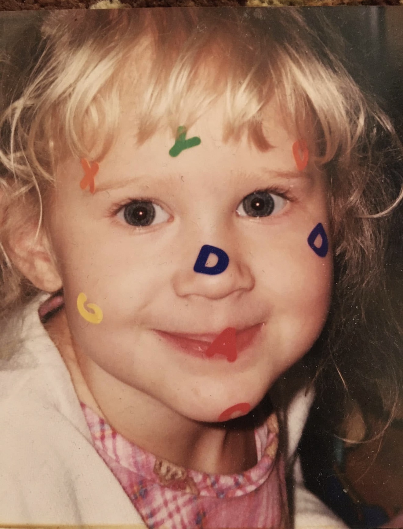 Child with curly blonde hair and big eyes, smiling with colorful marker drawings on face, wearing a pink and white checkered shirt.