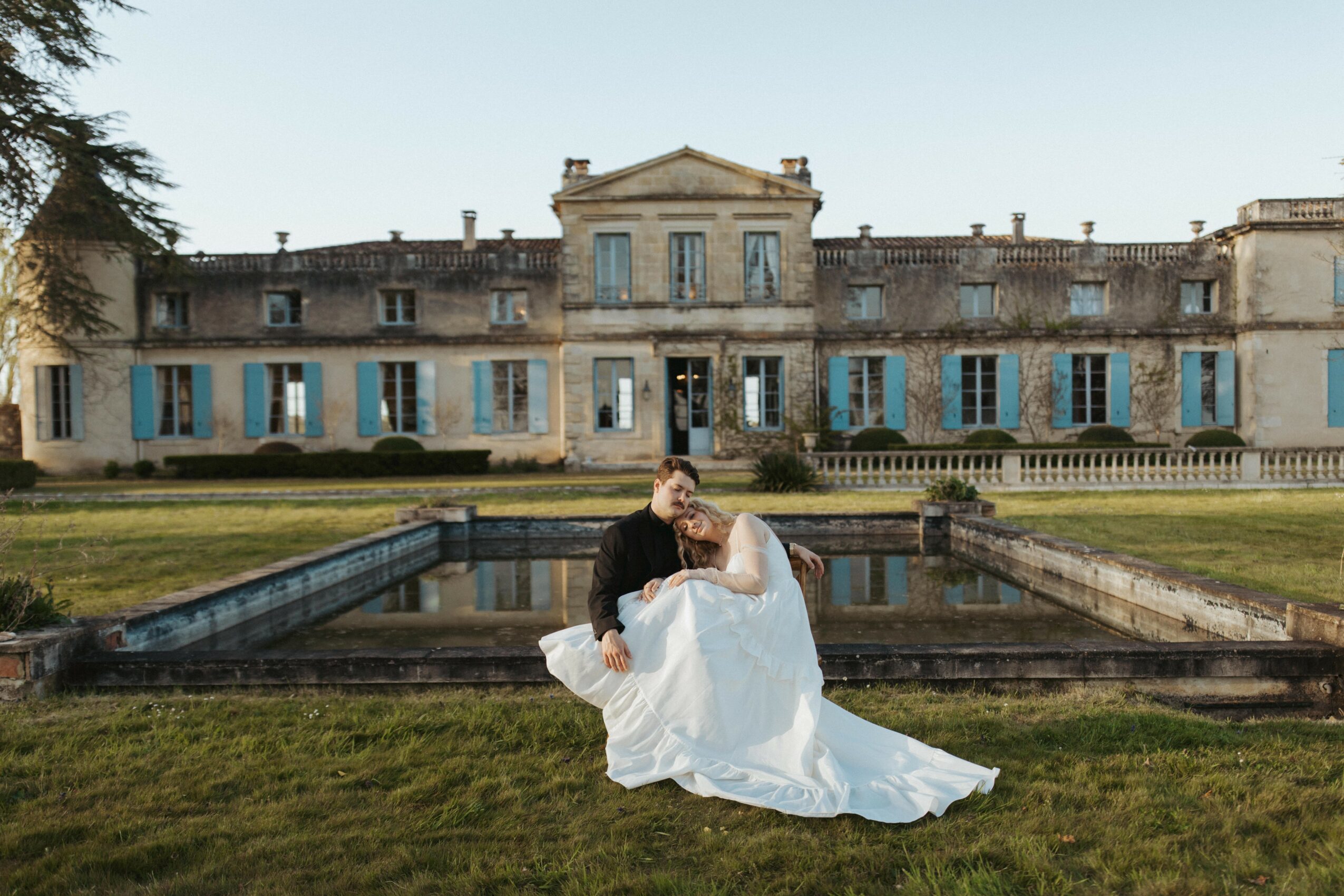 Couple in wedding attire sitting on grass in front of a large historic building with blue shutters.