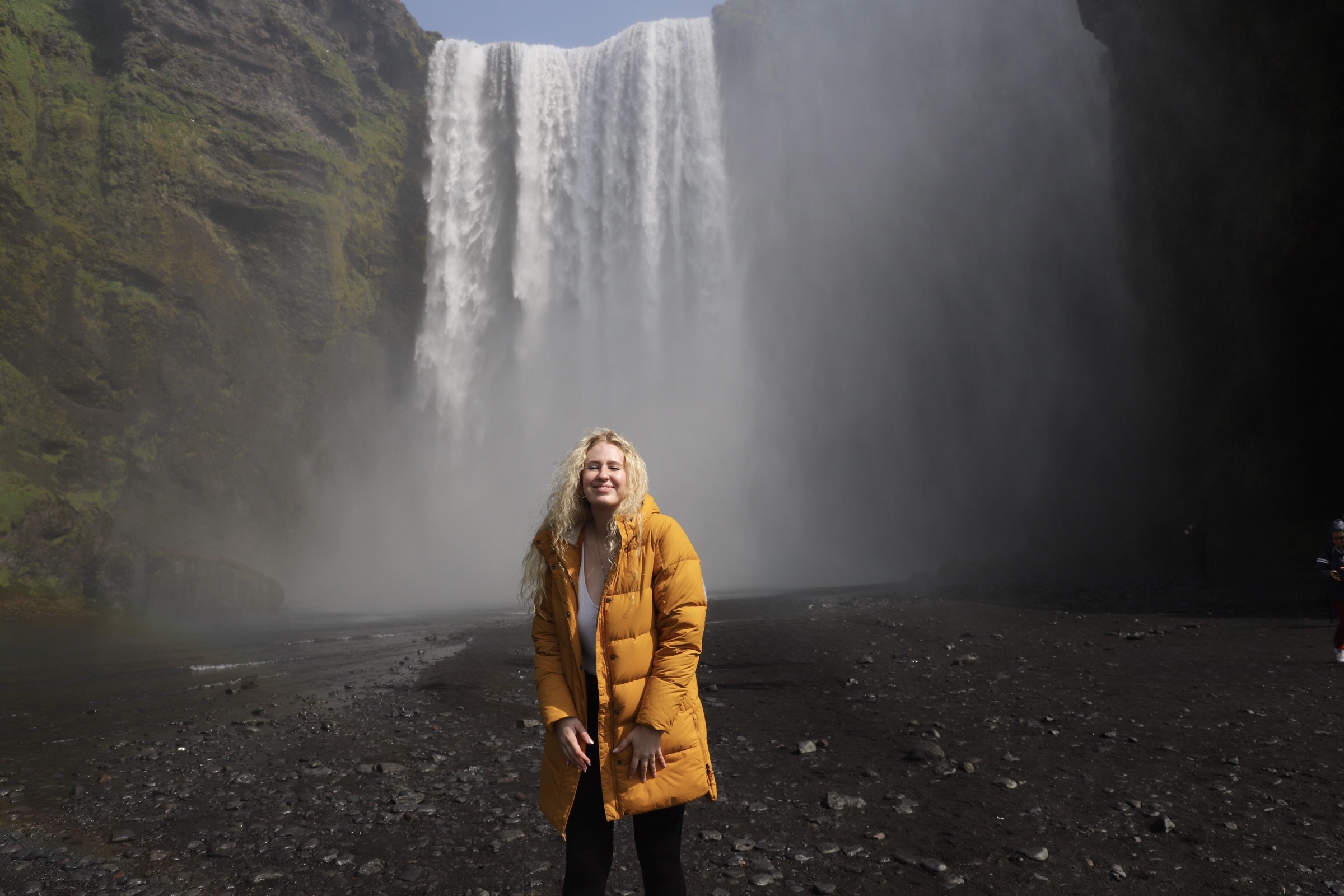 Person in yellow coat standing in front of a large waterfall with mist and rocky ground.