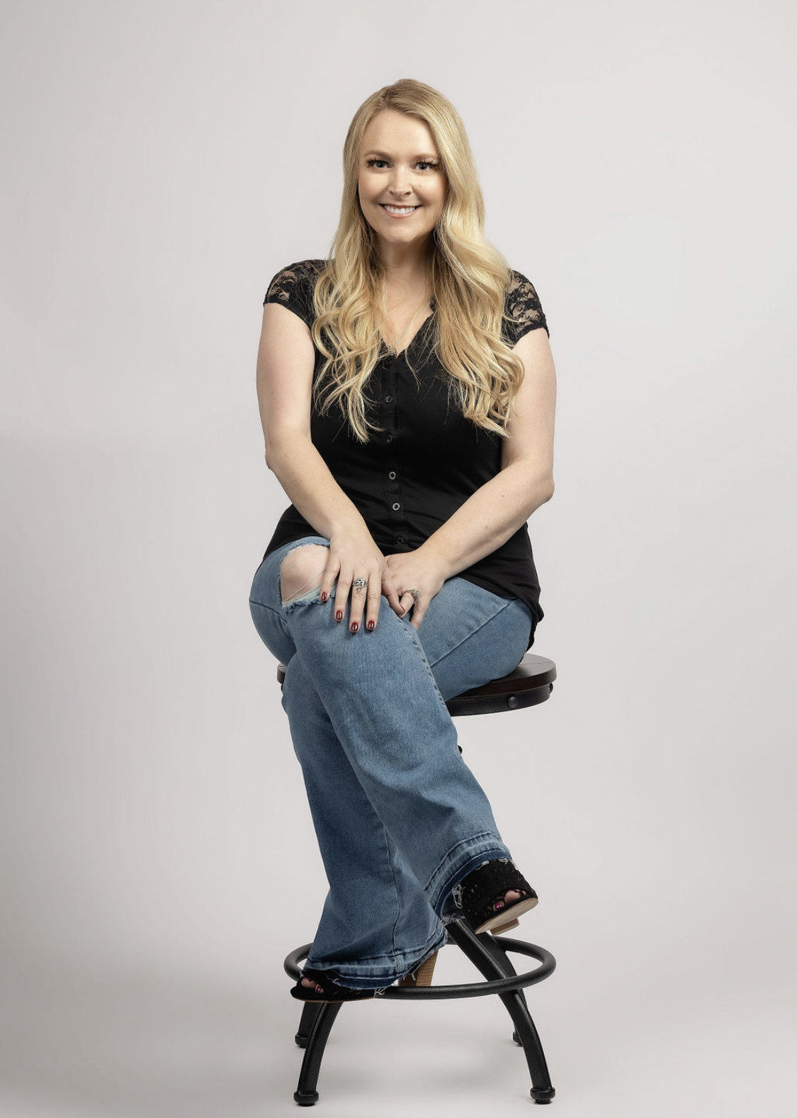 Woman with long blonde hair sitting on a stool, smiling, wearing a black top and blue jeans, against a plain background.
