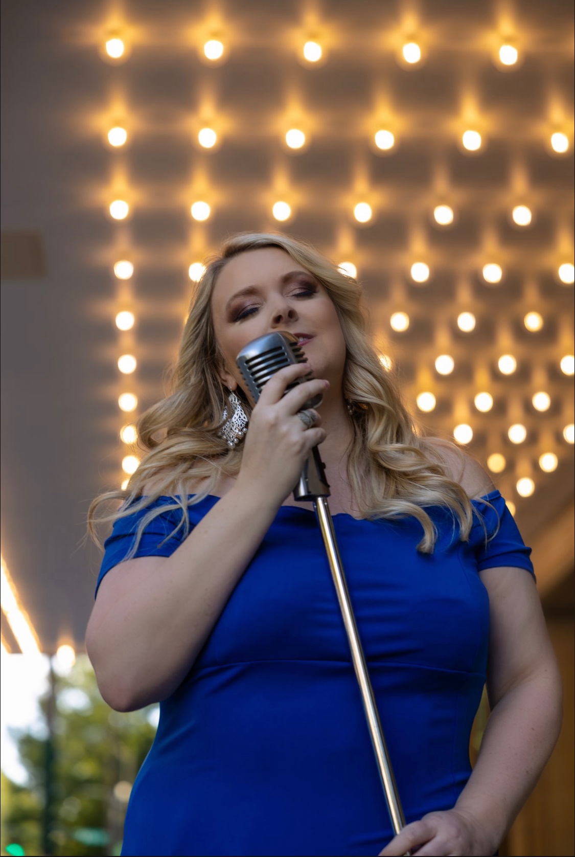 A woman with long blonde hair singing into a vintage microphone on stage, illuminated by bright lights overhead.