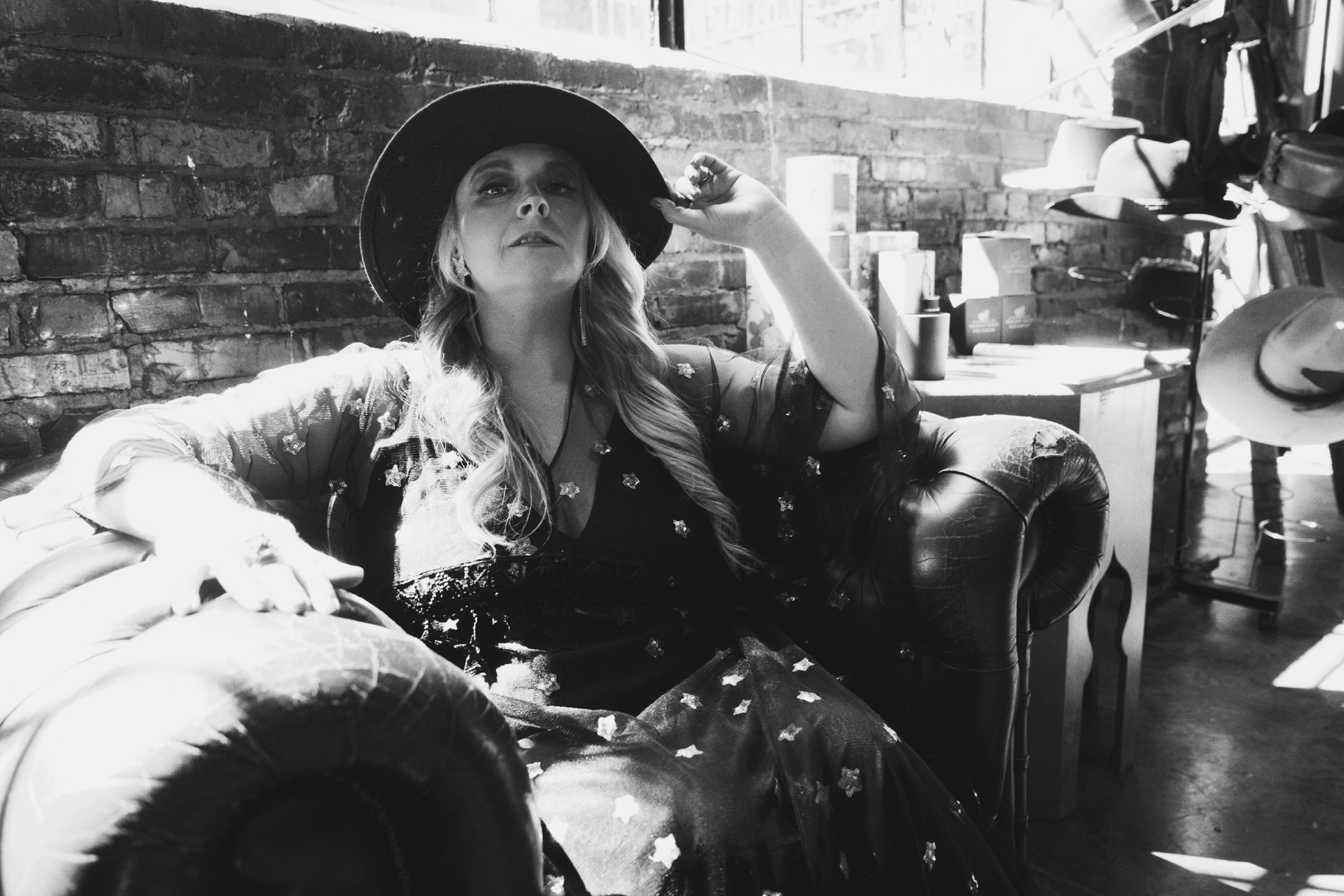 Woman sitting on a sofa wearing a hat, with a brick wall background and various objects around her.