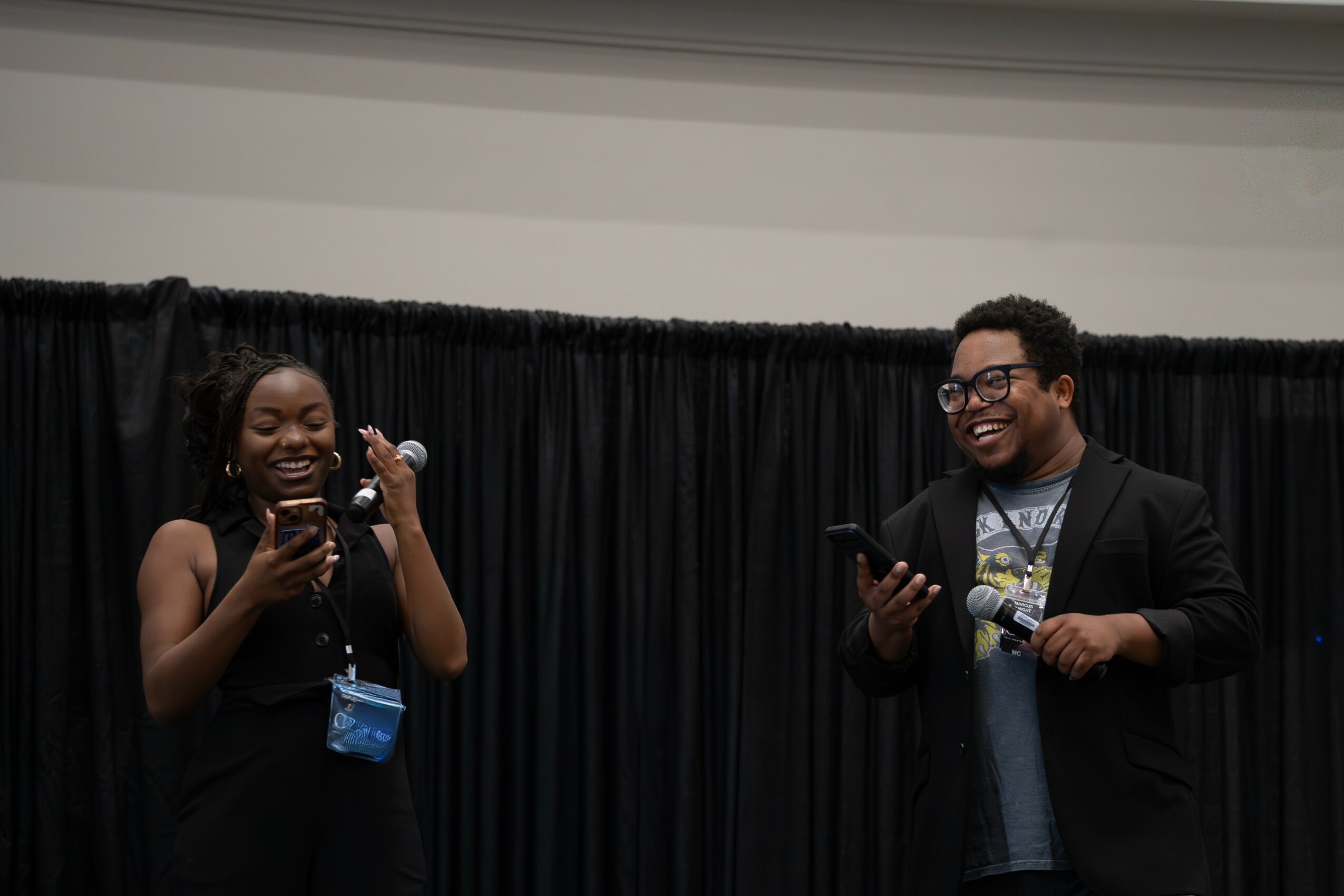 Two people standing in front of a black curtain, smiling and looking at their phones.