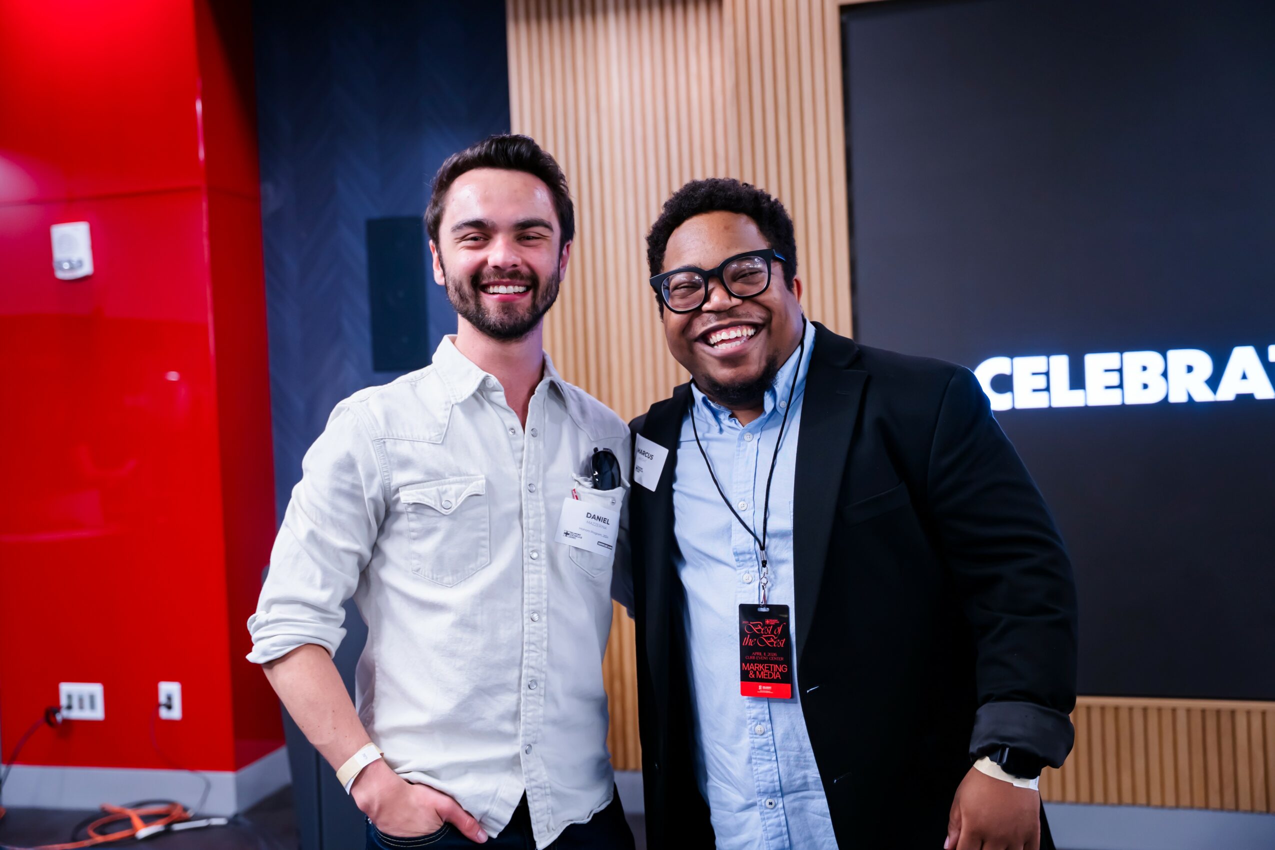 Two men smiling and standing together indoors, one in a white shirt and the other in a black blazer, with a screen behind them.