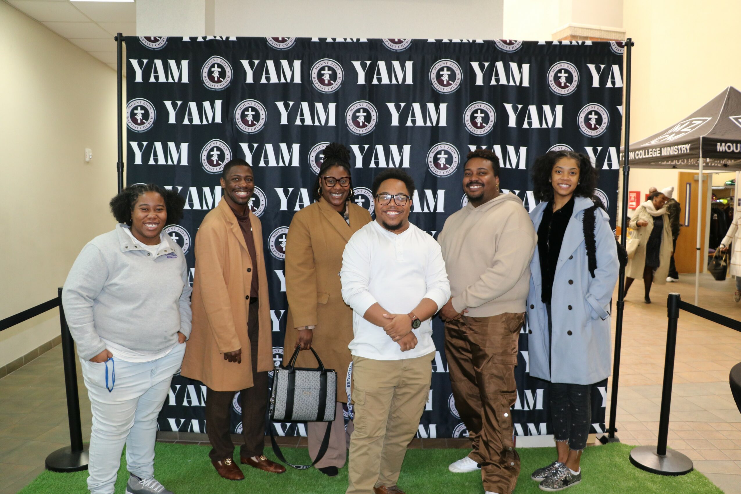 Group of six people standing in front of a YAM branded backdrop, smiling, indoors, with a tent and other people in the background.