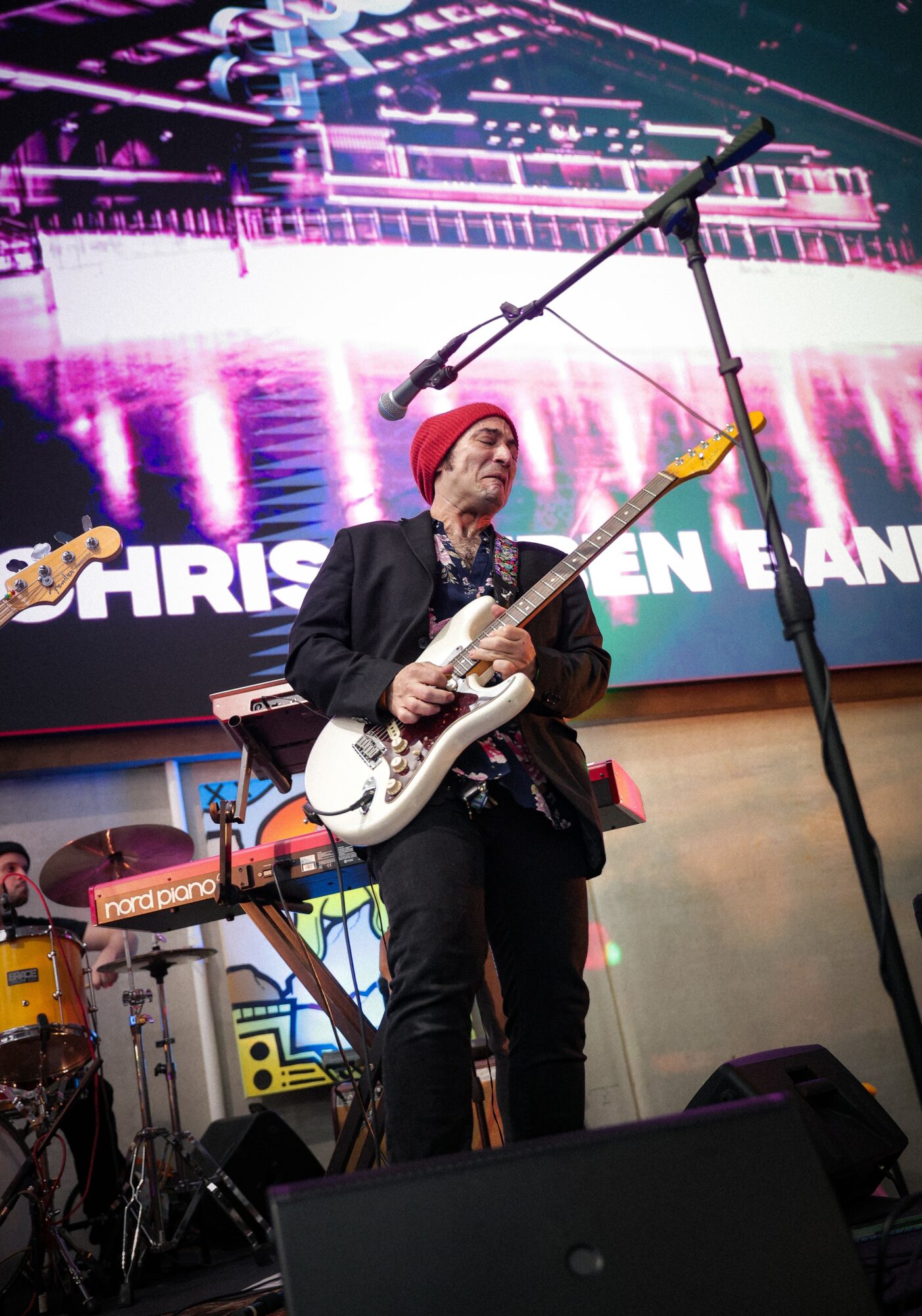 Musician with red hair and black clothing playing an electric guitar on stage, with a large colorful screen behind.