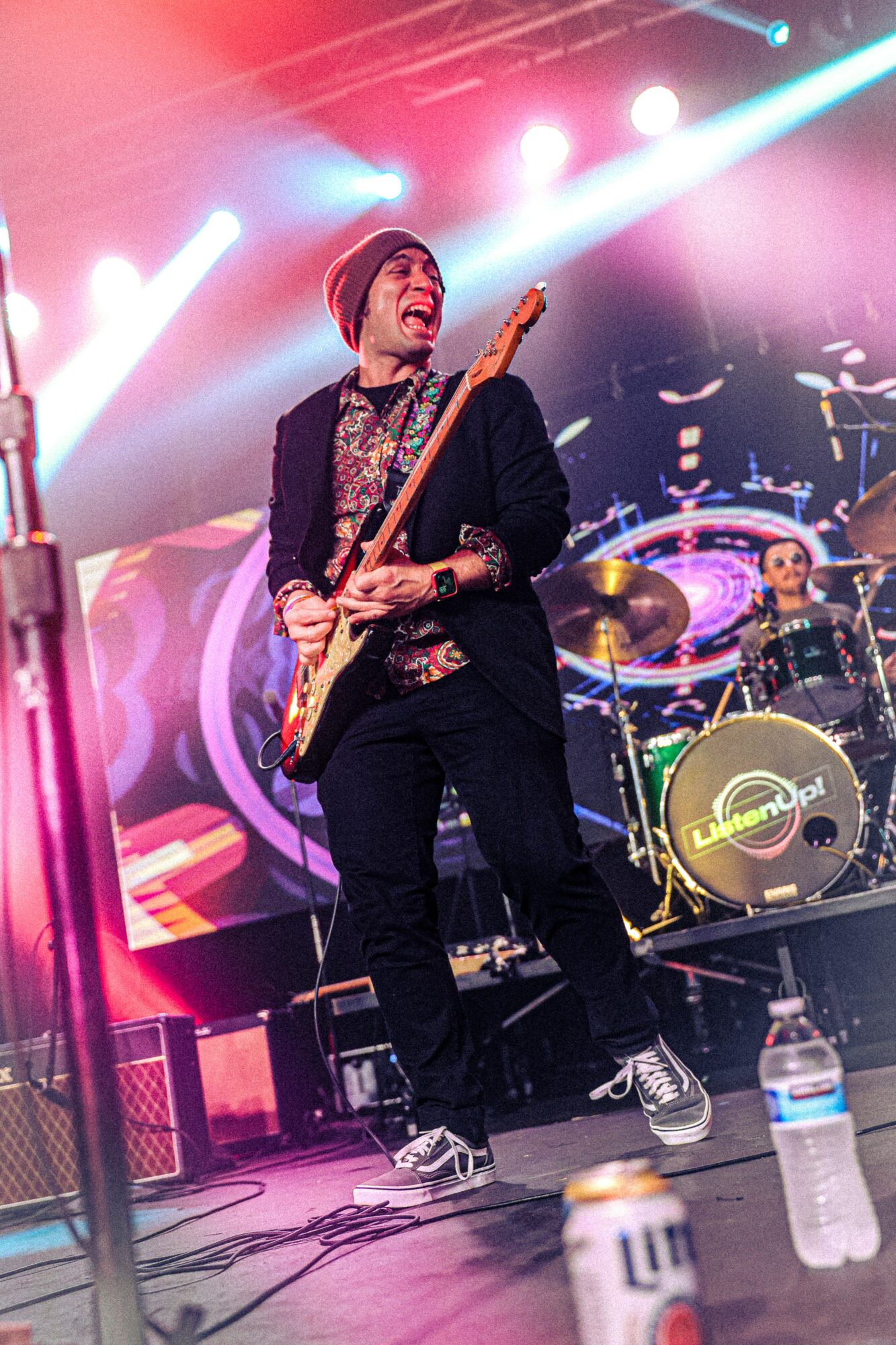 Musician playing electric guitar on stage with colorful lights and drum set in background.