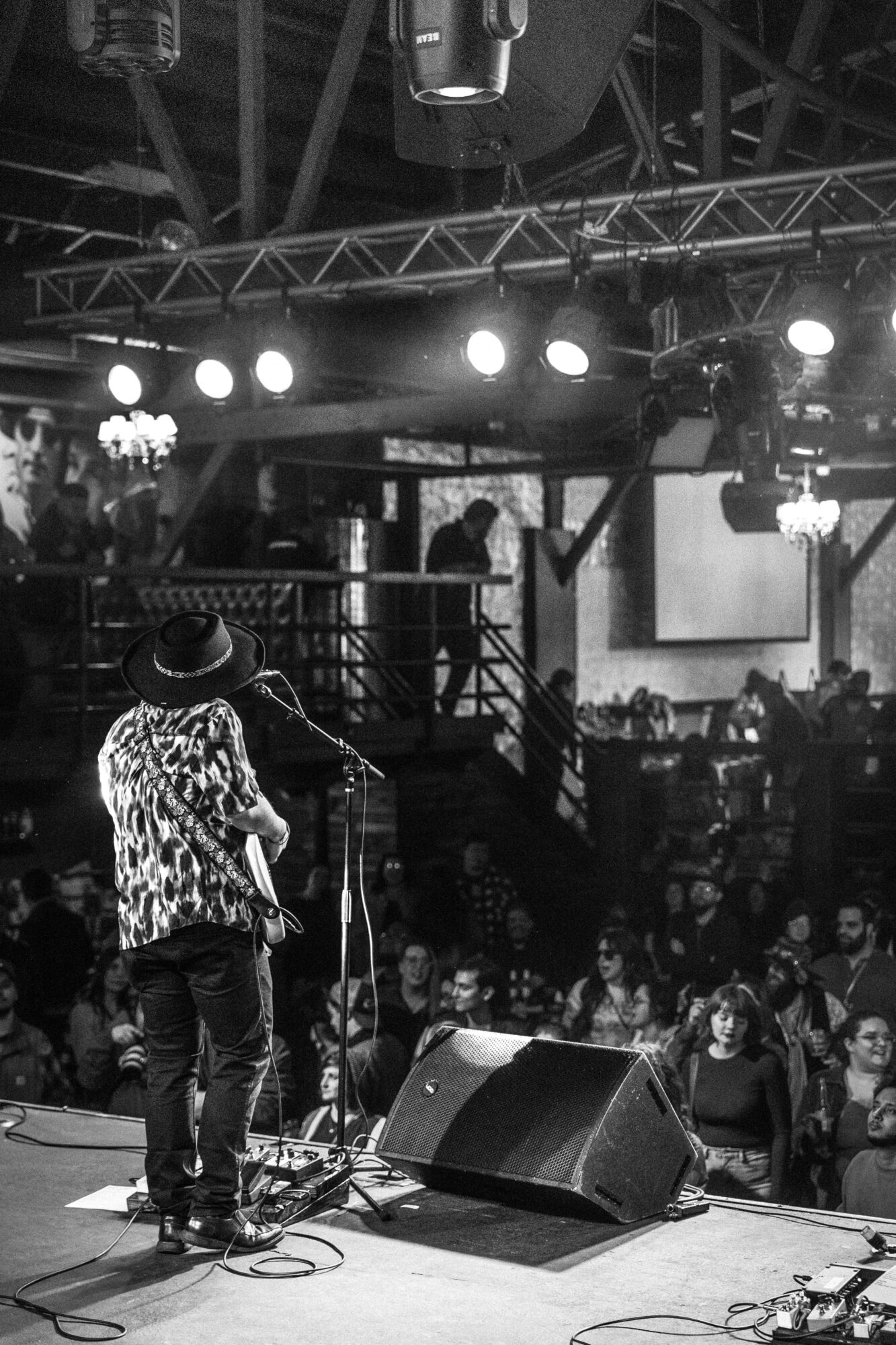 Performer with a hat and patterned shirt on stage, microphone, and audience in a dark indoor venue.