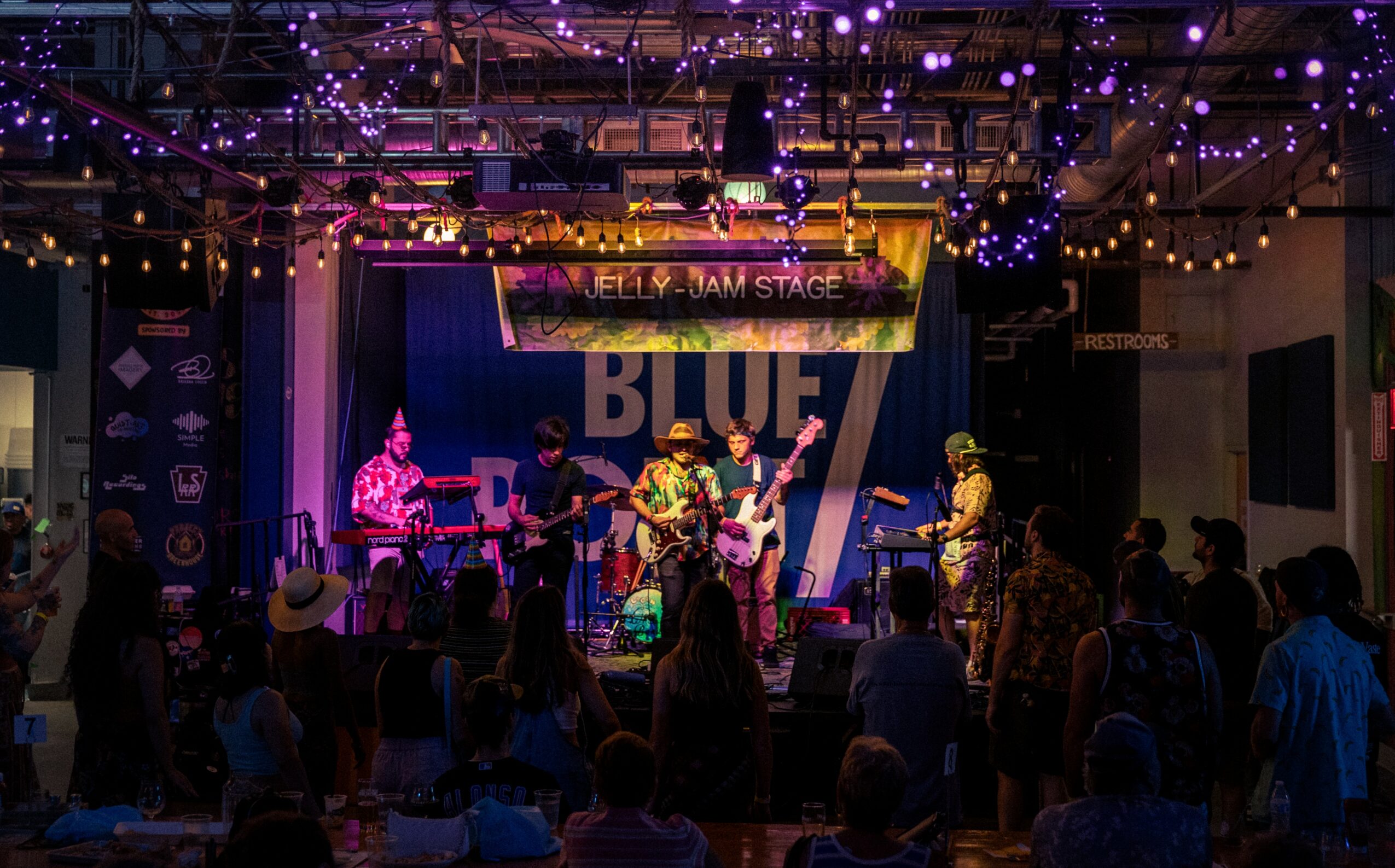 Musicians perform on stage with purple and yellow lighting, audience watching in a decorated venue with string lights.