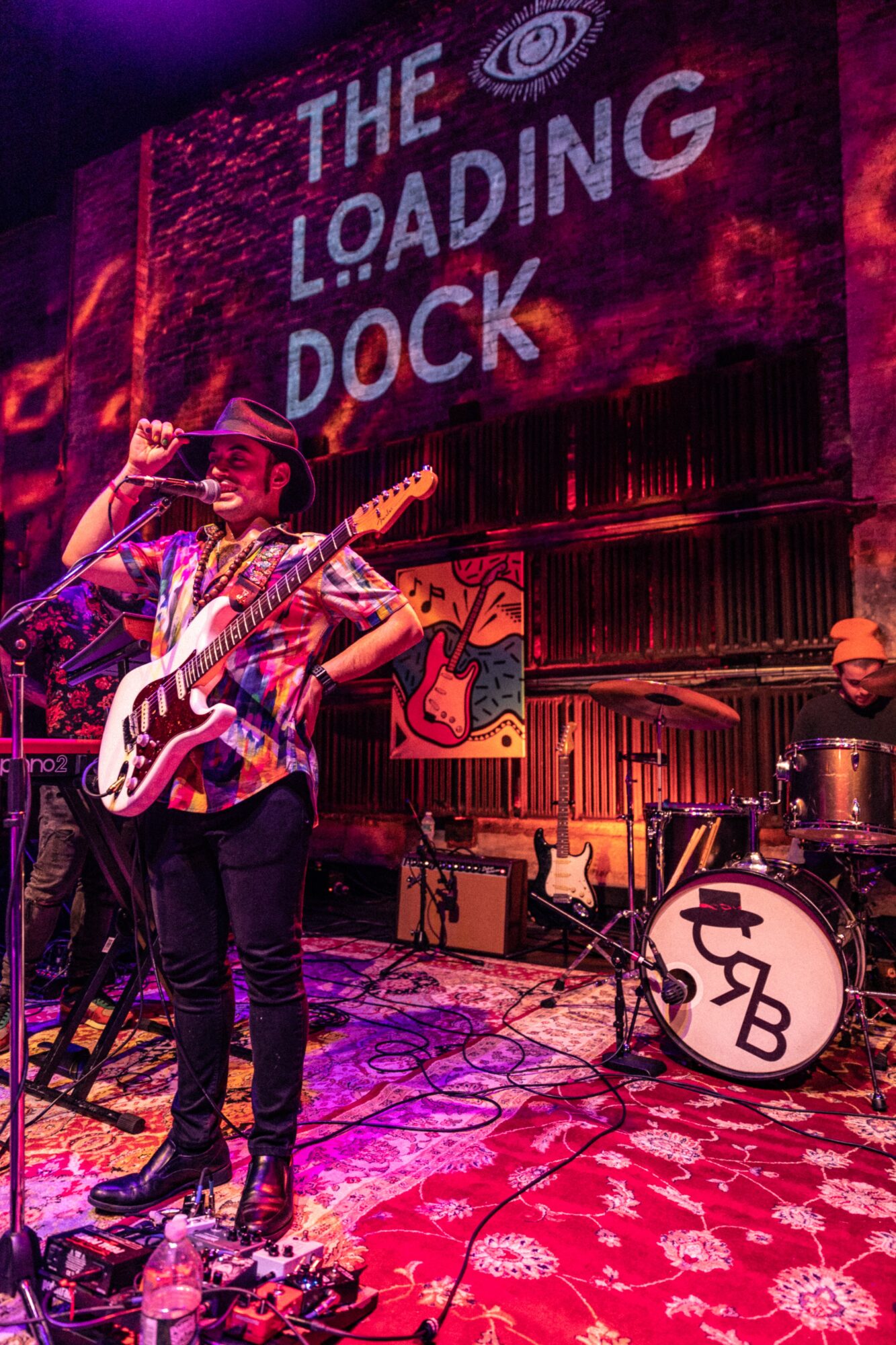 Musician singing into microphone with guitar, band playing drums in background, stage with colorful lighting, and a sign reading 'The Loading Dock'.