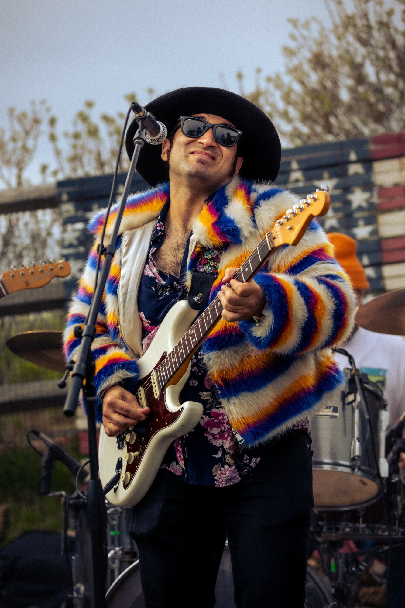 Musician wearing sunglasses and a colorful striped jacket playing an electric guitar outdoors.