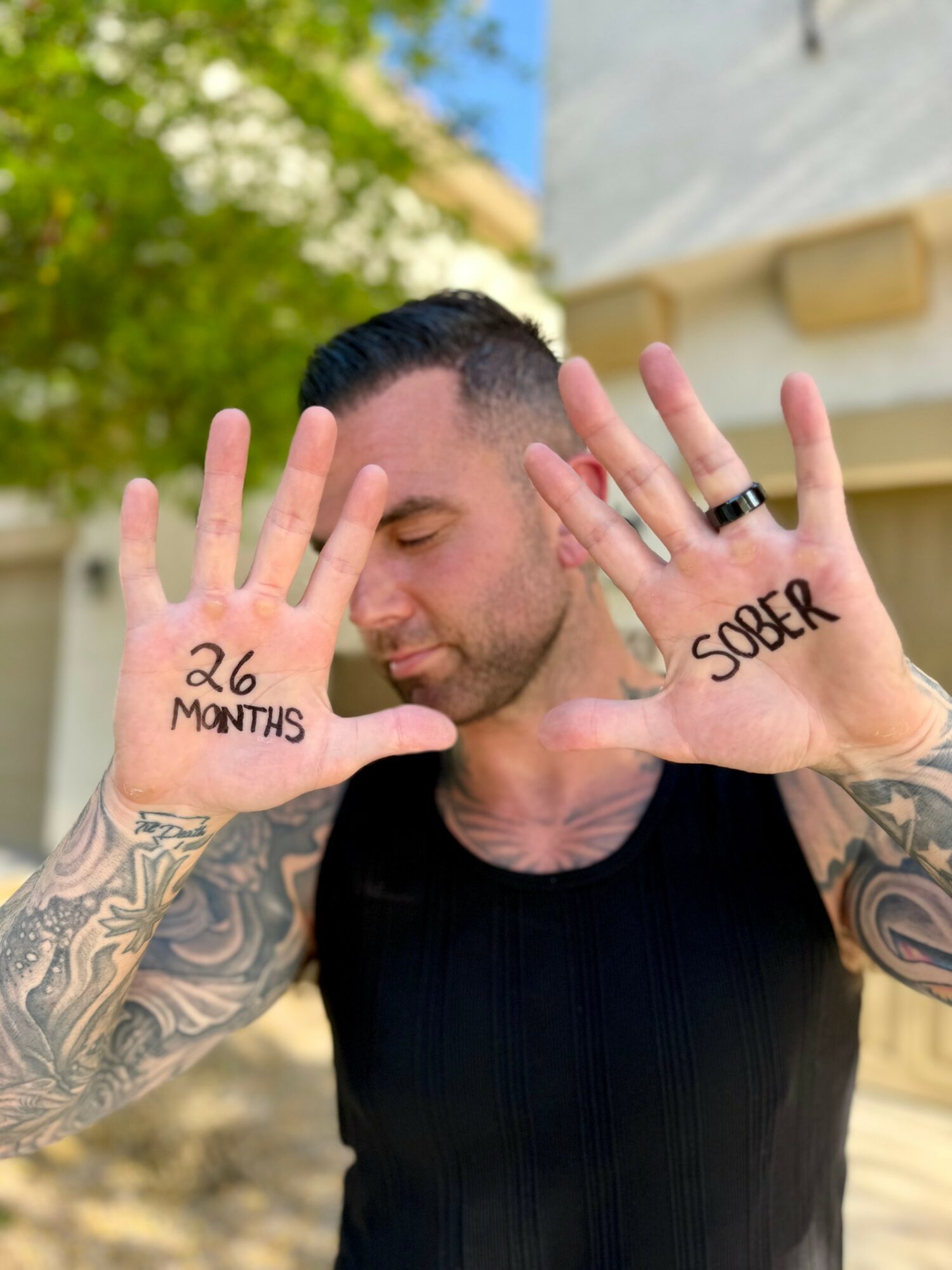 Man with tattoos holding up hands with words '26 MONTHS' and 'SOBER' written on palms, outdoors.