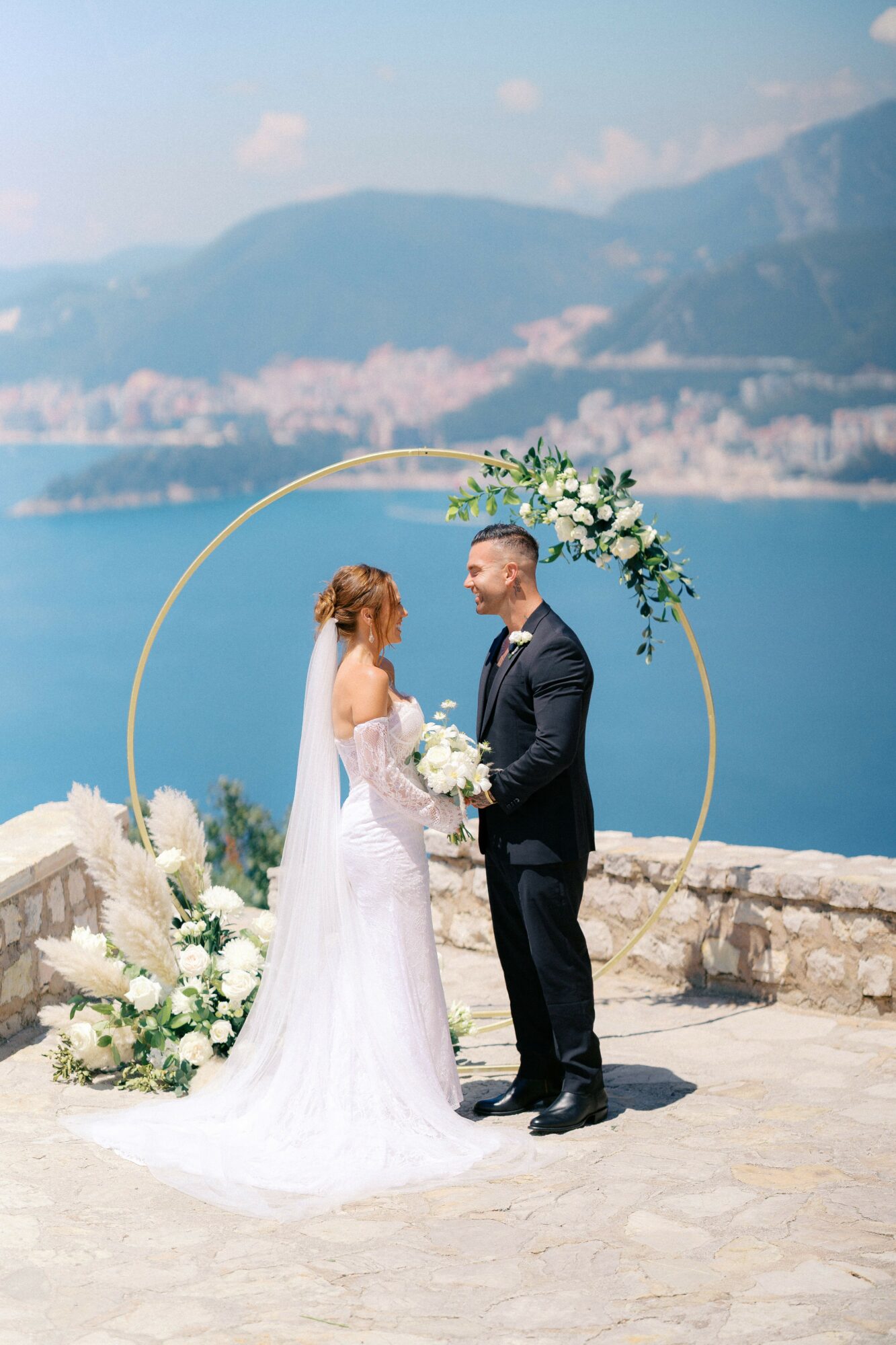 Bride and groom standing outdoors by a stone wall, holding hands, with a lake and mountains in the background, decorated with flowers and a circular arch.