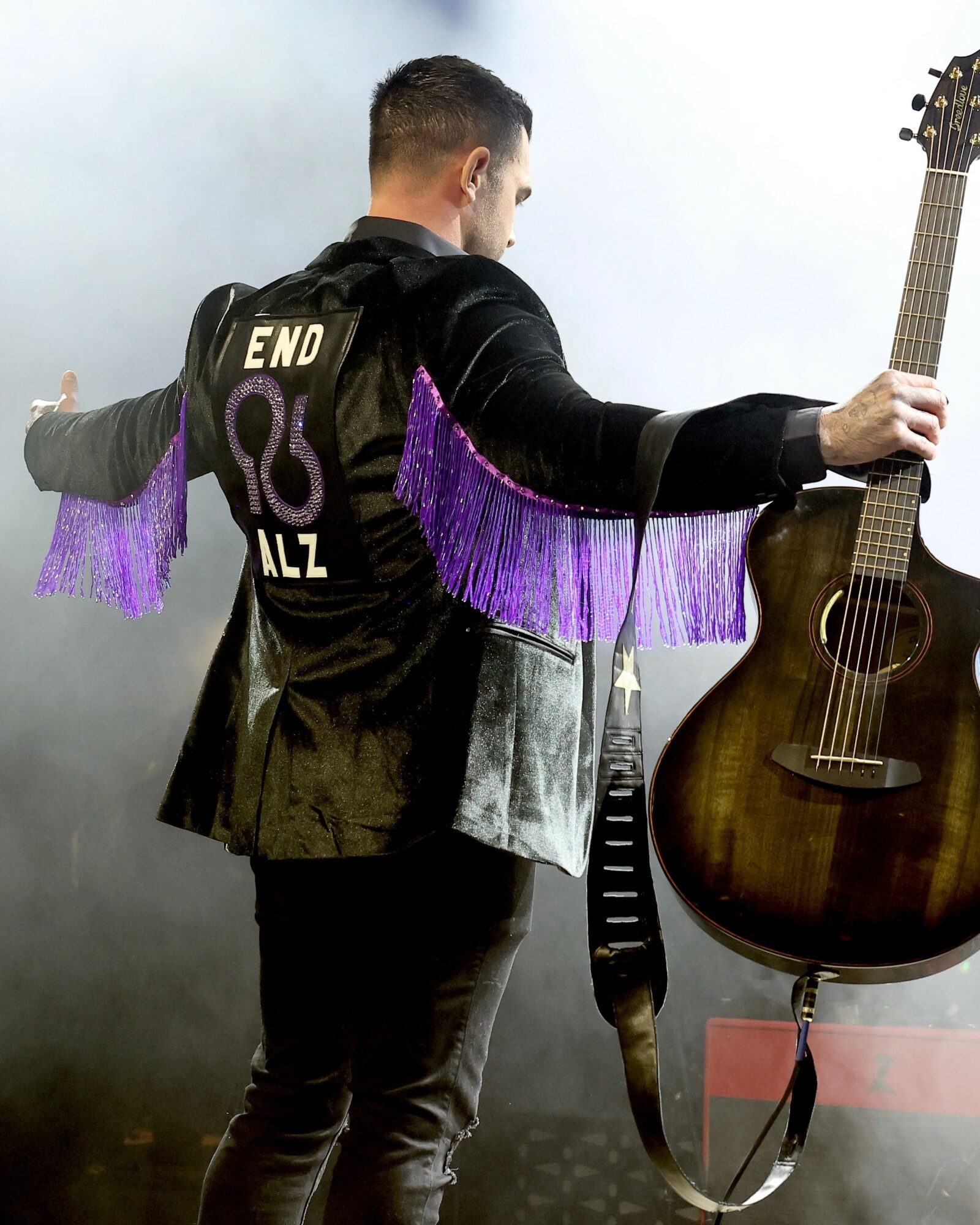 Person with arms outstretched holding a guitar, facing away, wearing a black jacket with purple fringe details, against a smoky background.
