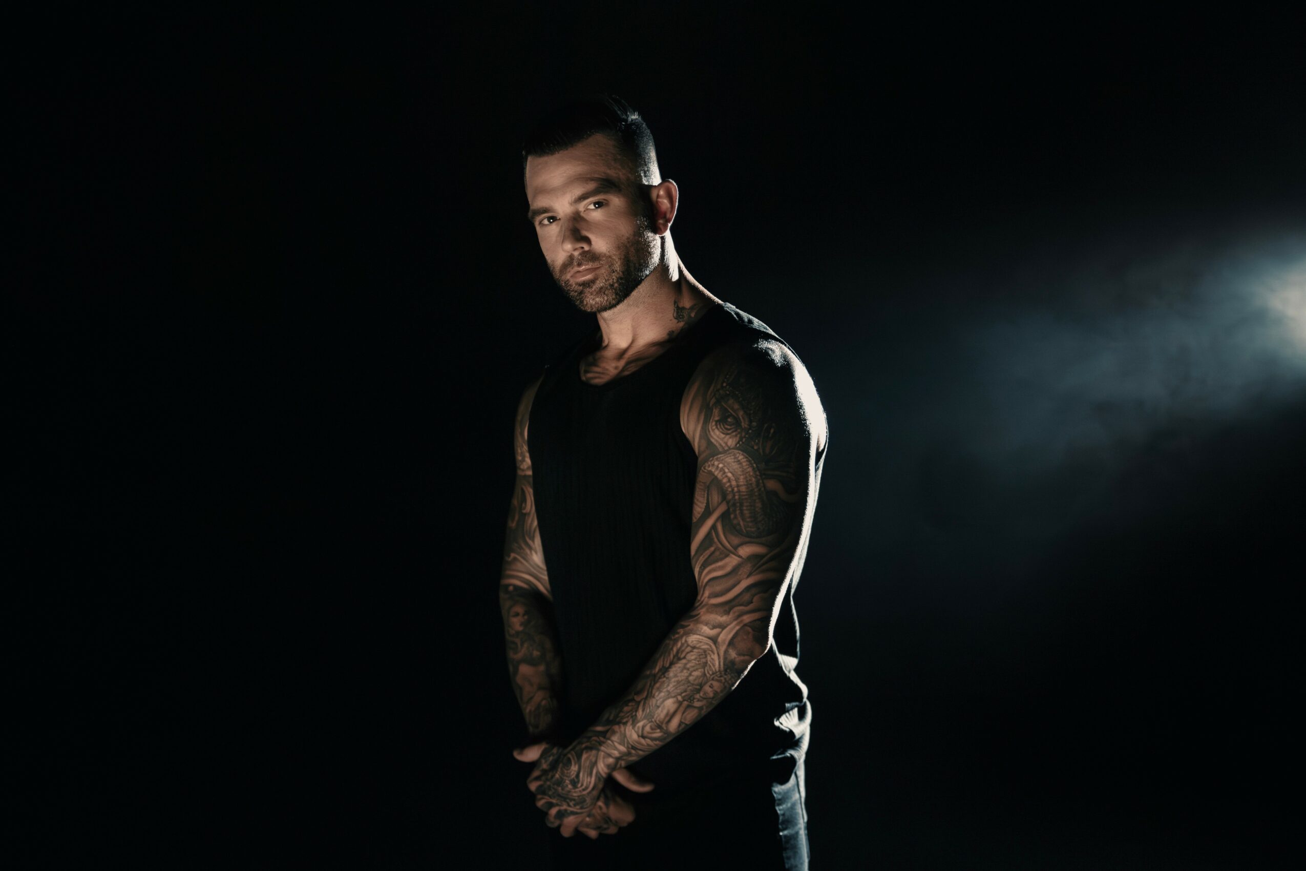 A man with tattoos on his arms stands in a dark setting with a light source behind him, looking at the camera.
