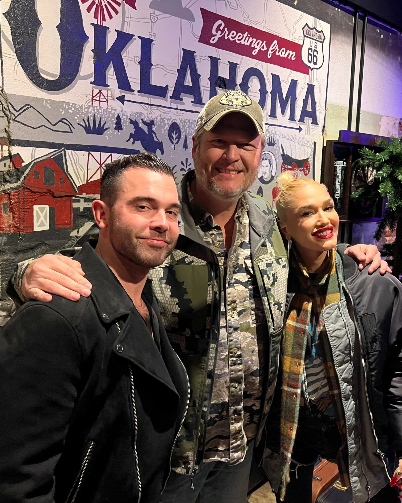 Three people smiling, standing together in front of a Oklahoma-themed backdrop with a Route 66 sign and greeting message.