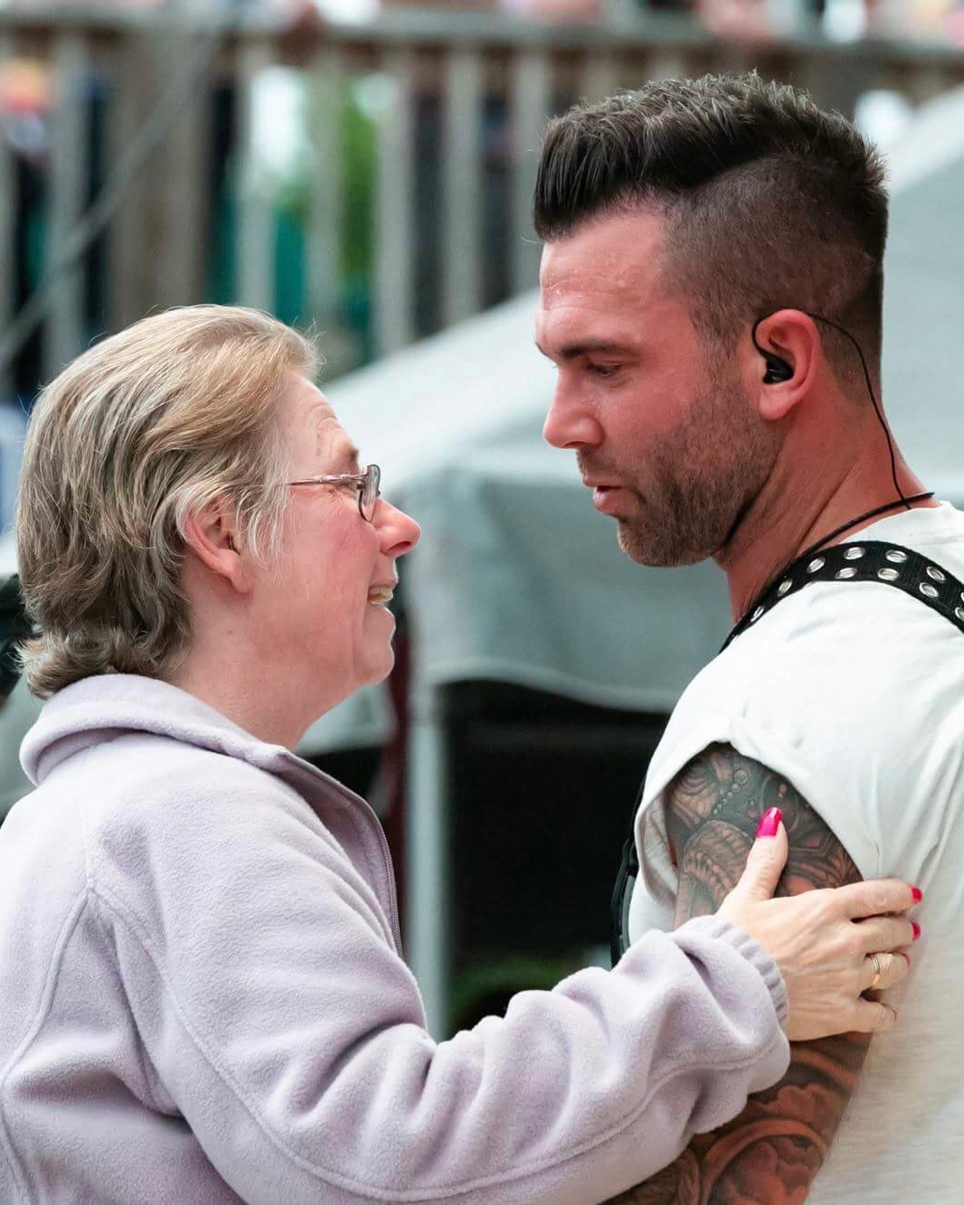 An elderly woman and a young man face each other closely, holding hands, outdoors with blurred background.